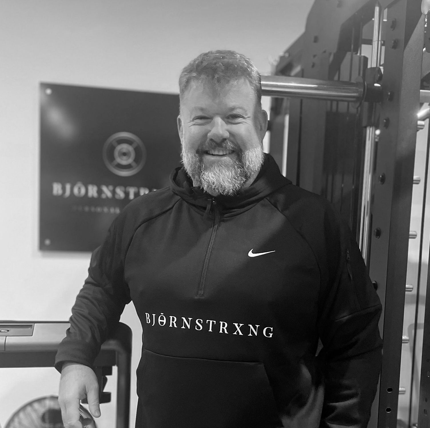 Man in a black hoodie with a beard, leaning on equipment in a gym. 