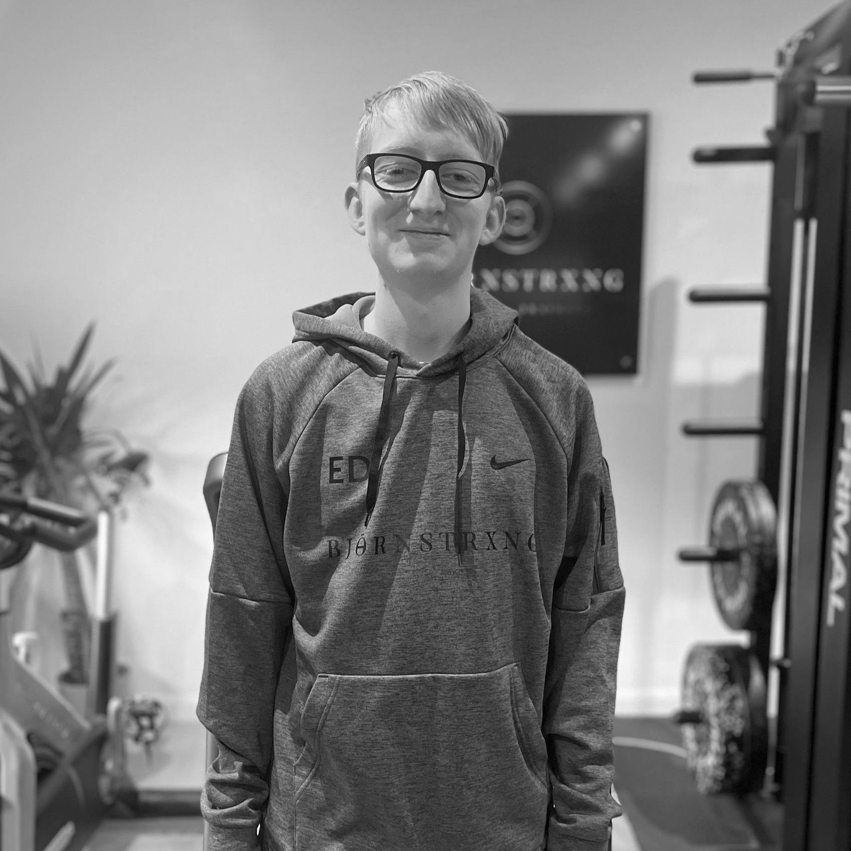 Man in a gray hoodie and glasses stands in a gym, posing in front of weightlifting equipment.
