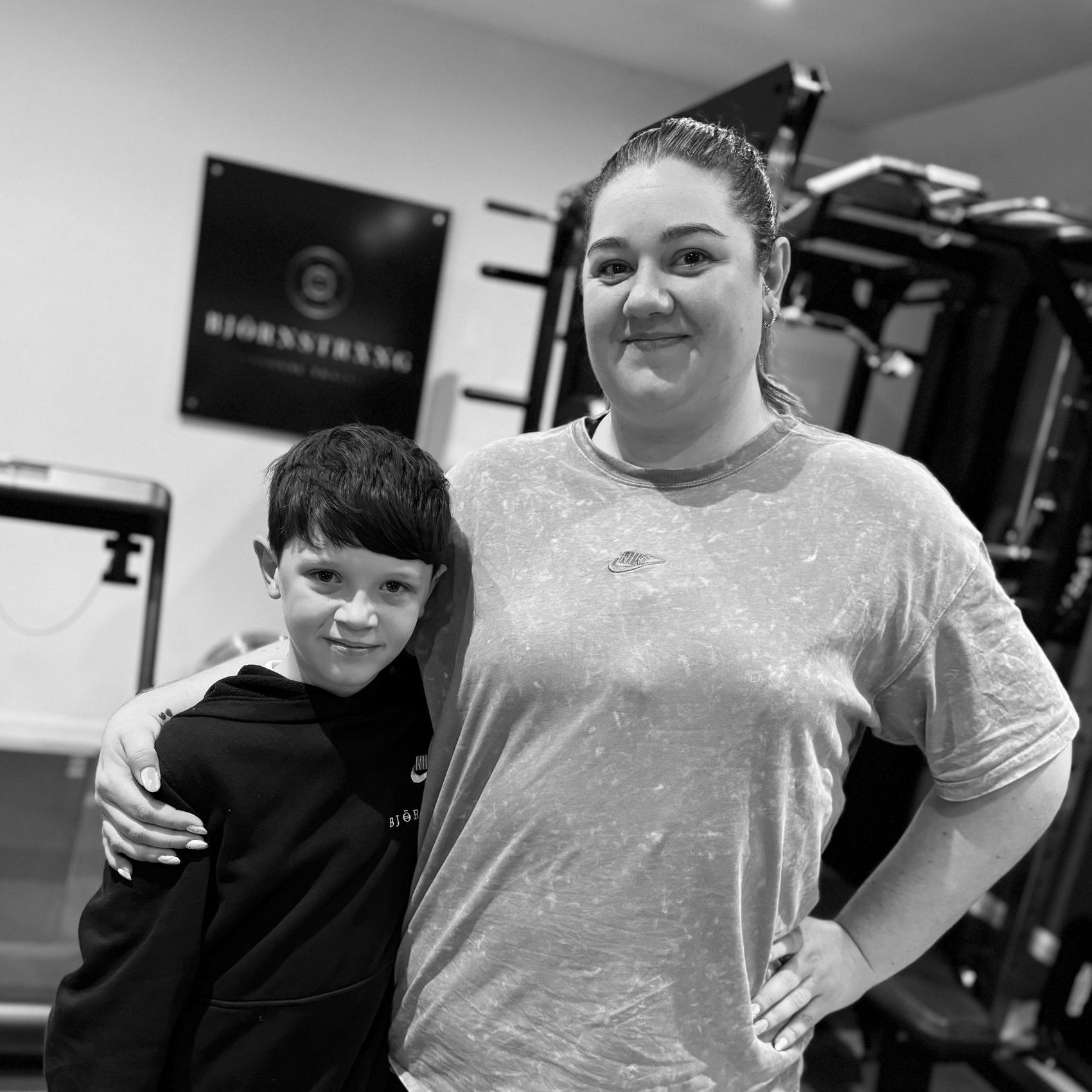 Woman with arm around a boy, smiling in a gym with equipment and a logo on the wall. Black and white photo.