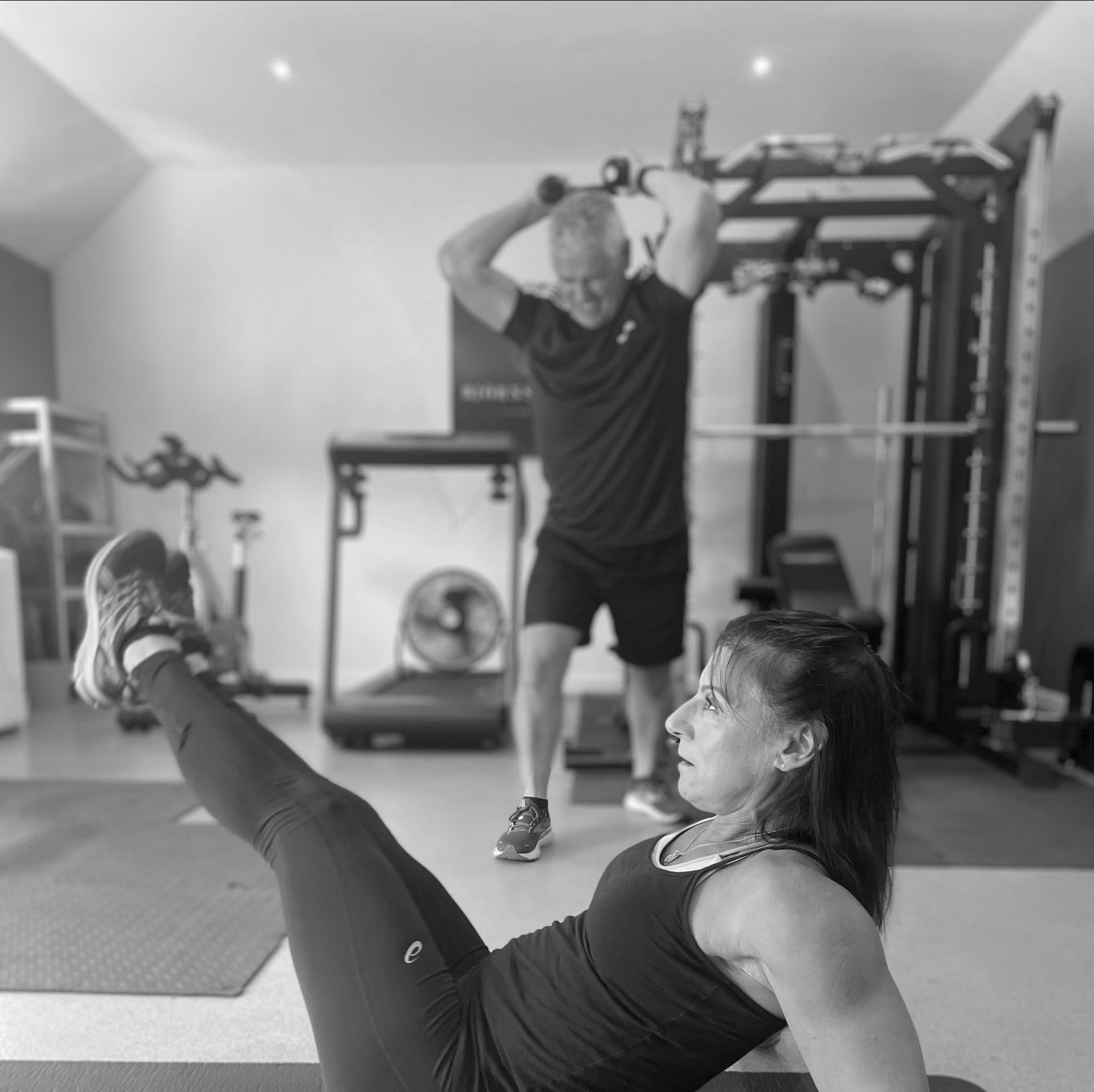 Woman doing core exercise while trainer exercises with weights in a gym.