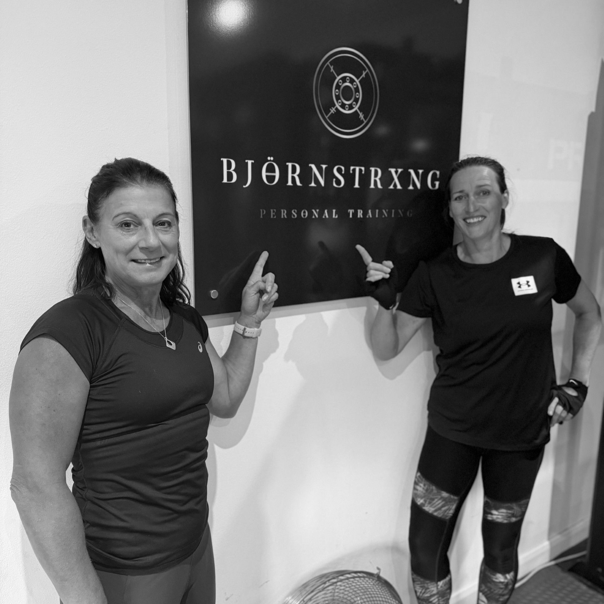 Four women in a gym, holding dumbbells, wearing white shirts with logos, smiling.