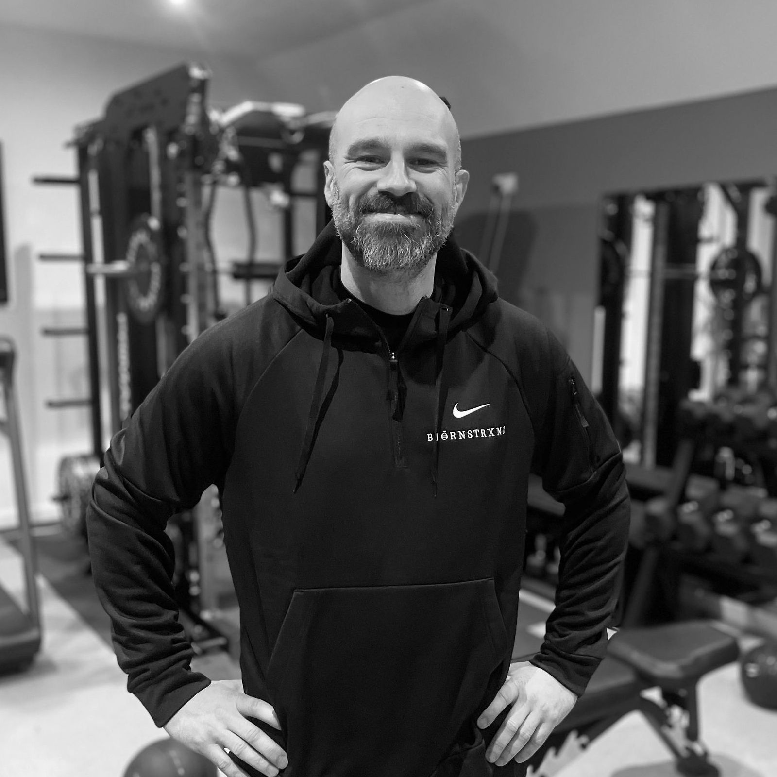 Man in black hoodie, hands on hips, smiling in a gym. Weight equipment in the background.