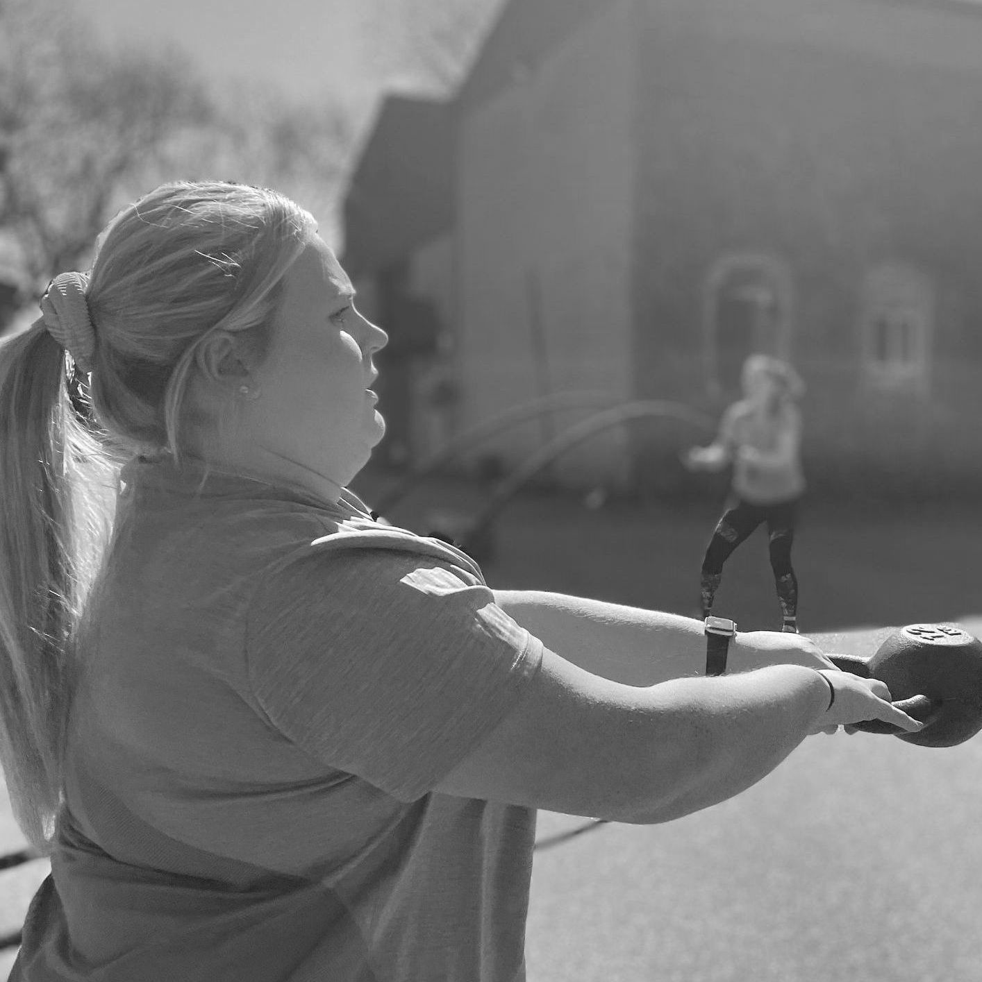 Woman holding a kettlebell, exercising outdoors. Another person is jumping rope in the background.