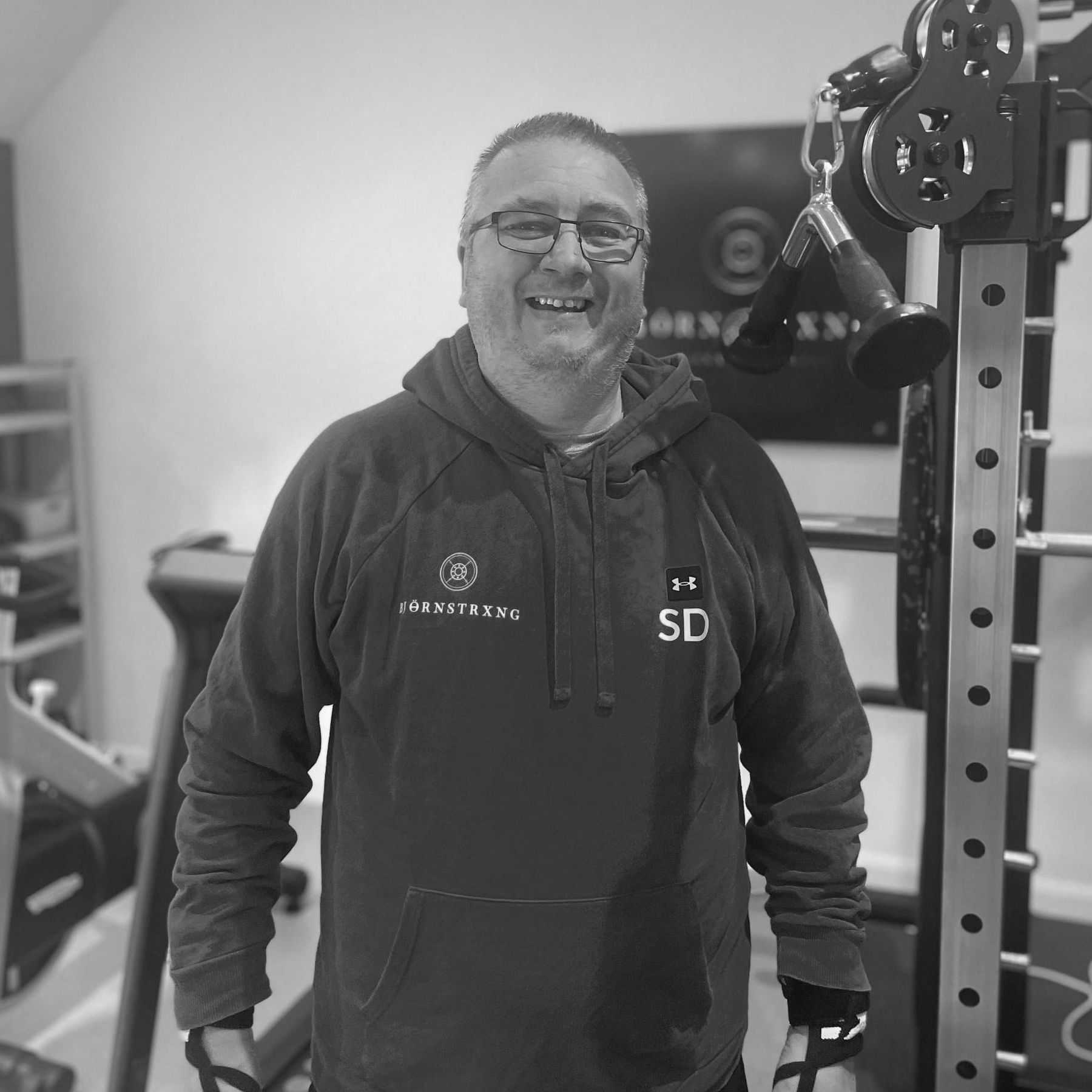 Man in hoodie smiles in home gym, posing near weight machine.