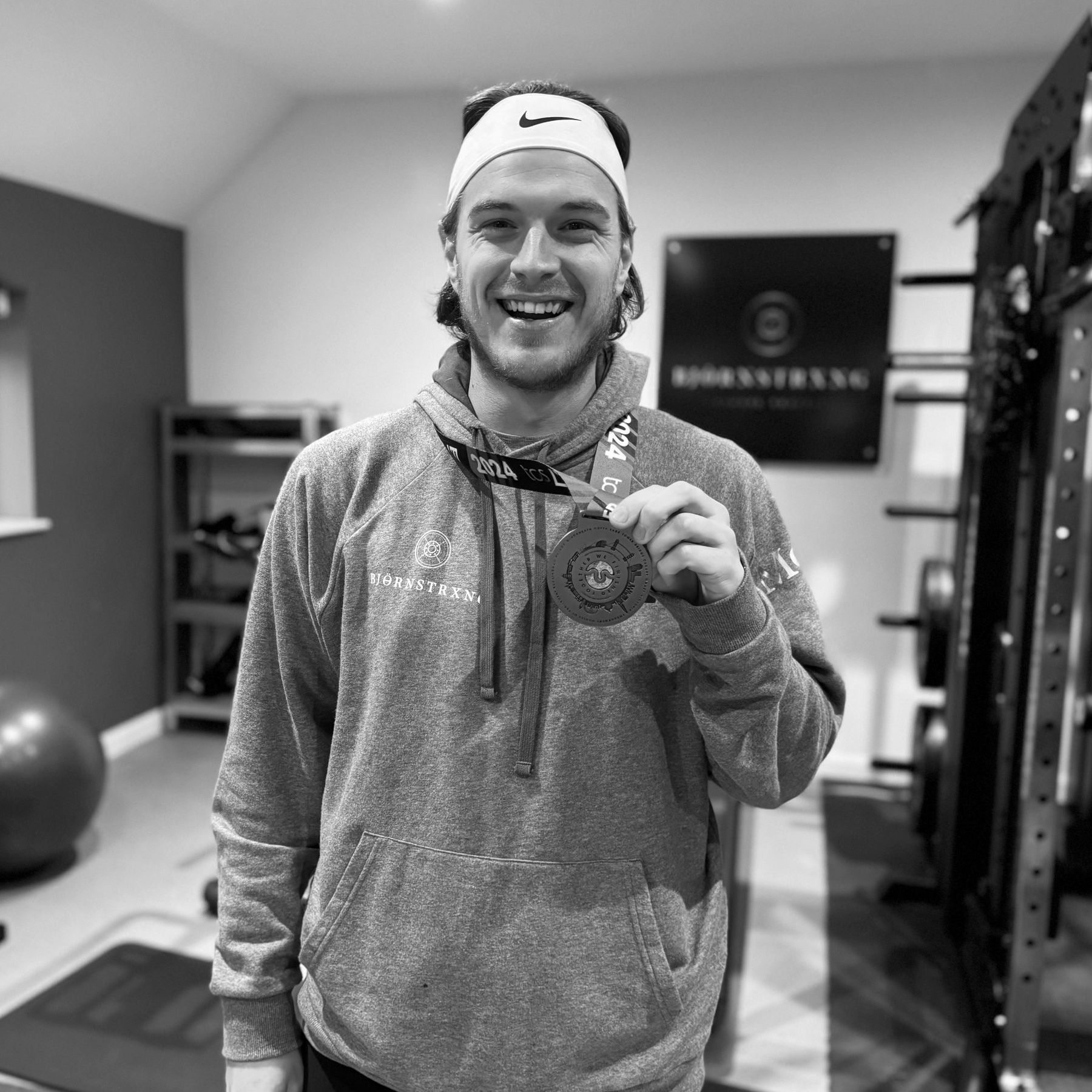 Man in a hoodie and headband smiles, holding a medal in a gym setting.
