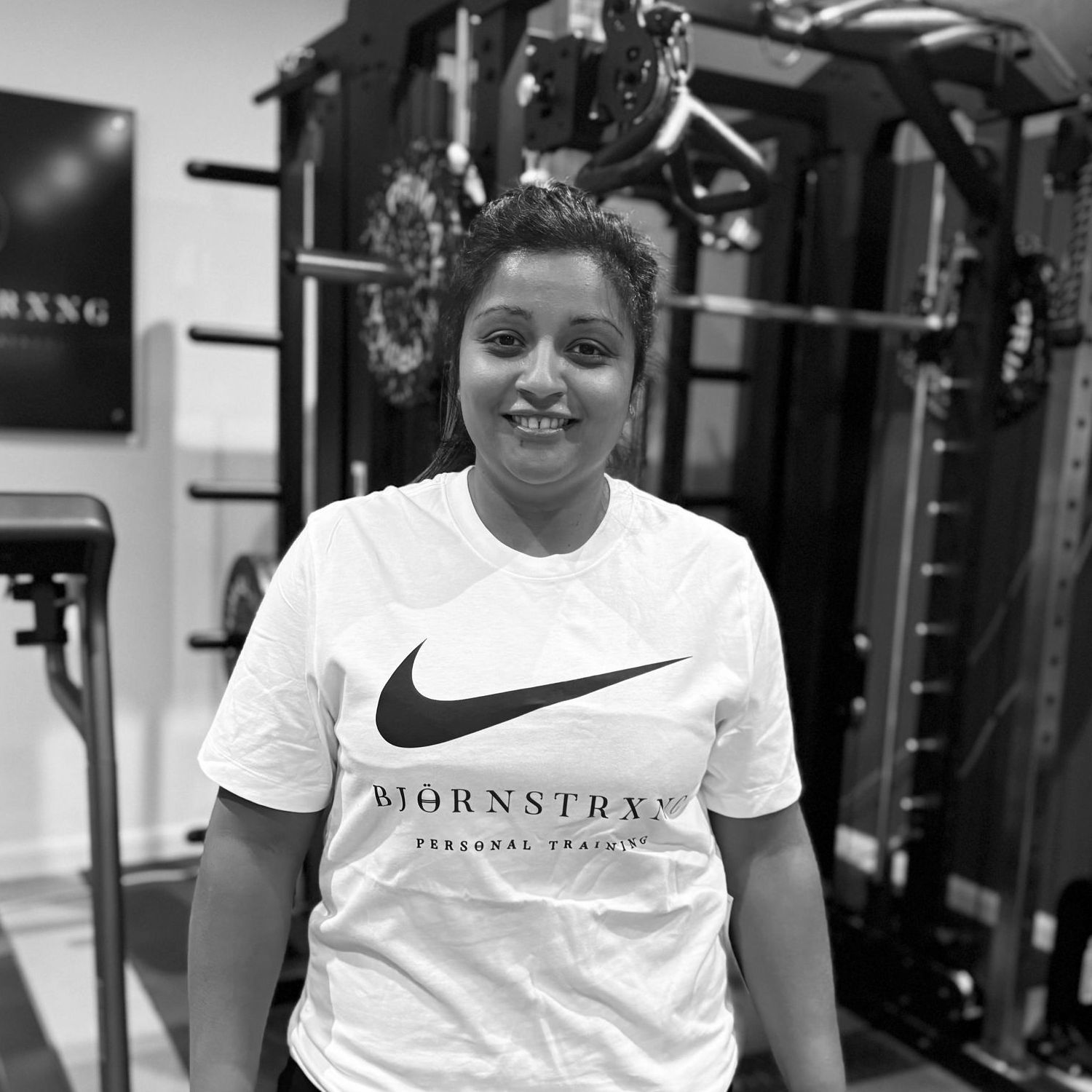 Woman smiling in a gym, wearing a Nike t-shirt, standing in front of exercise equipment.