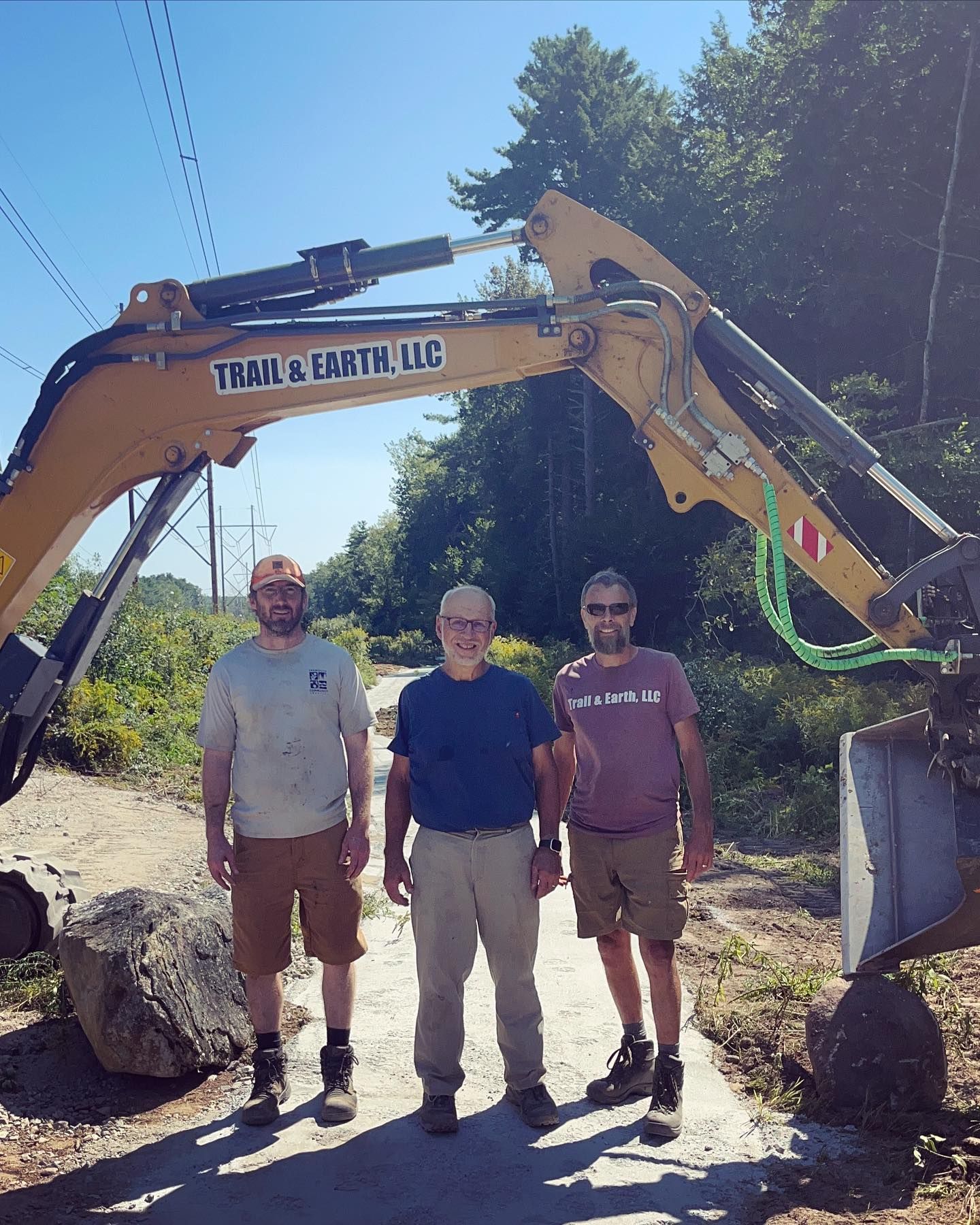 Three men stand by an excavator. Trail & Earth LLC logo visible. Trees and utility lines in the background.