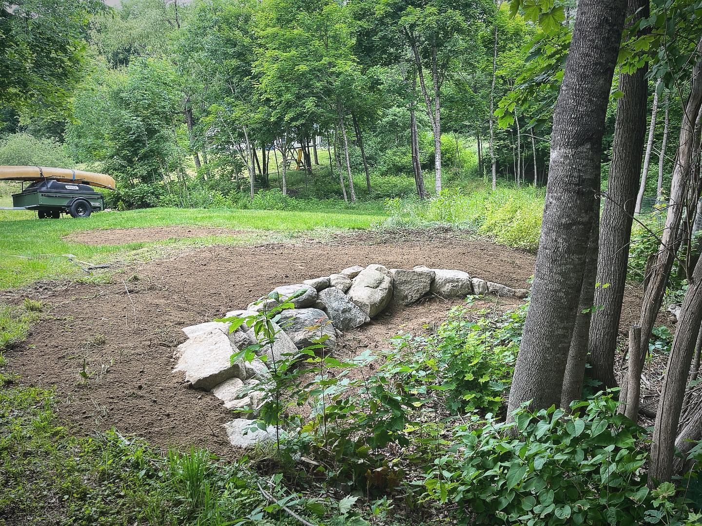 Newly tilled garden bed with a stone retaining wall, set among trees and green grass.