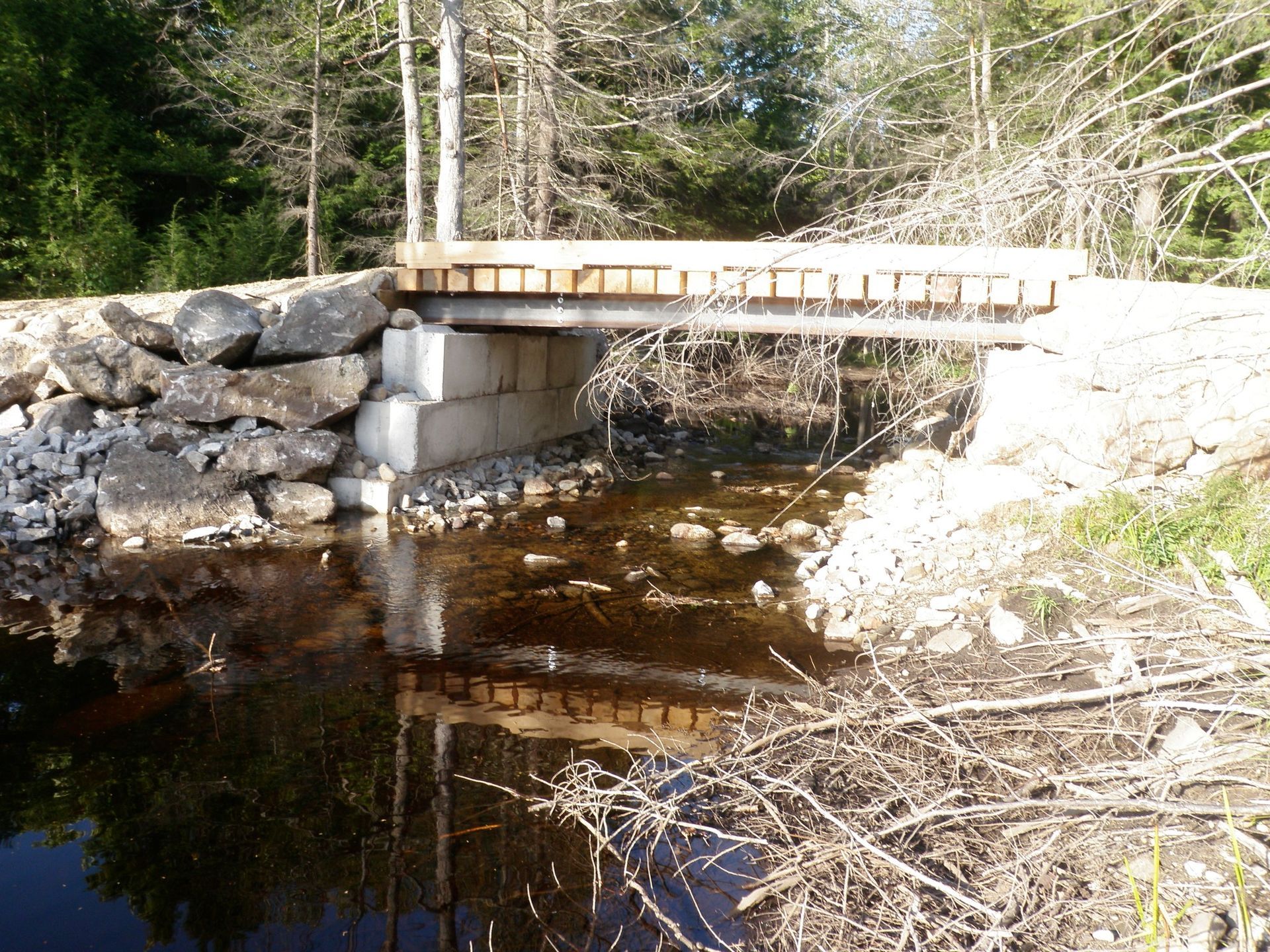 Small wooden bridge over a stream, with stone and concrete supports, surrounded by trees.