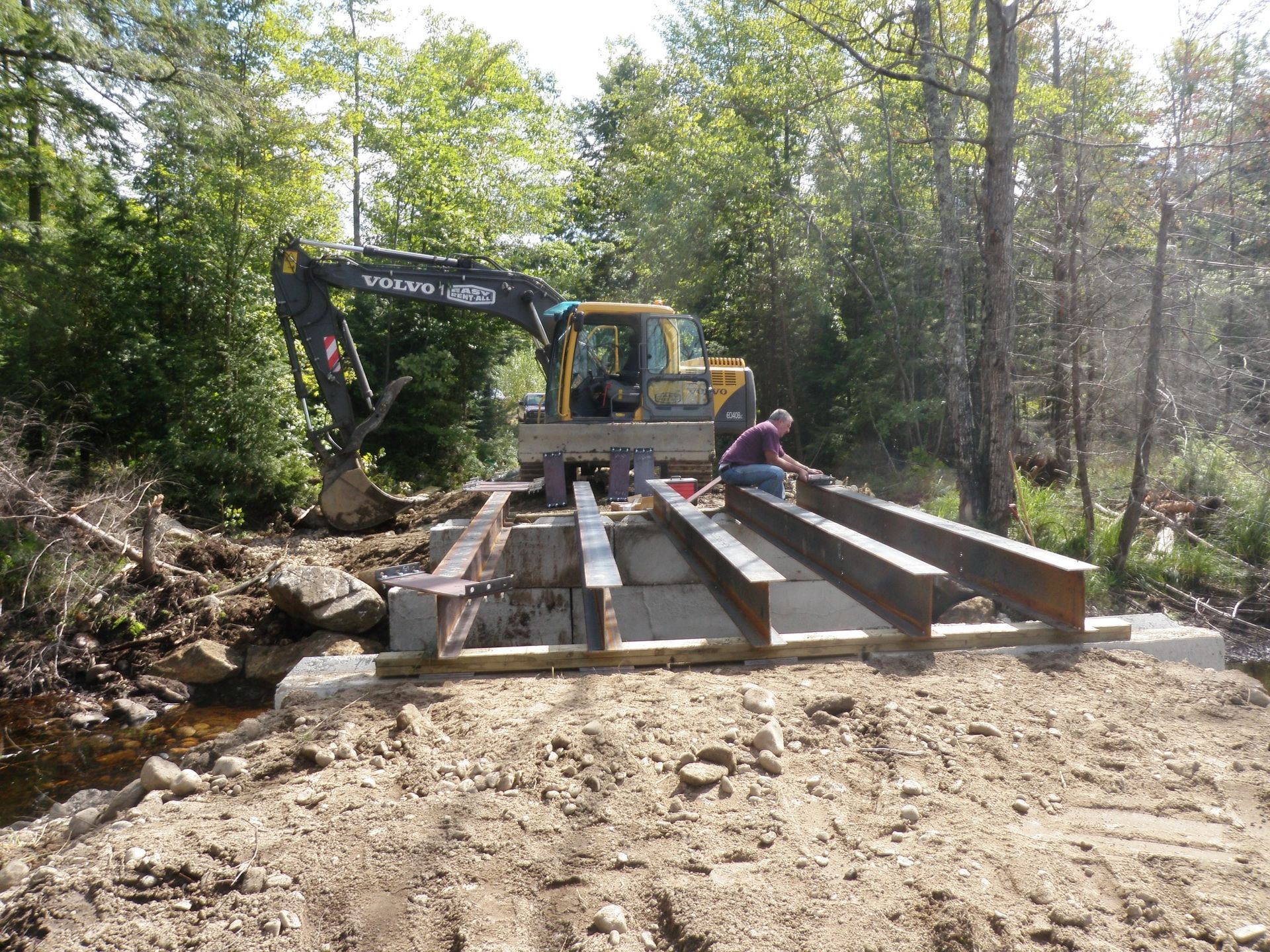 Construction site: excavator and person working on bridge over a creek in a wooded area.