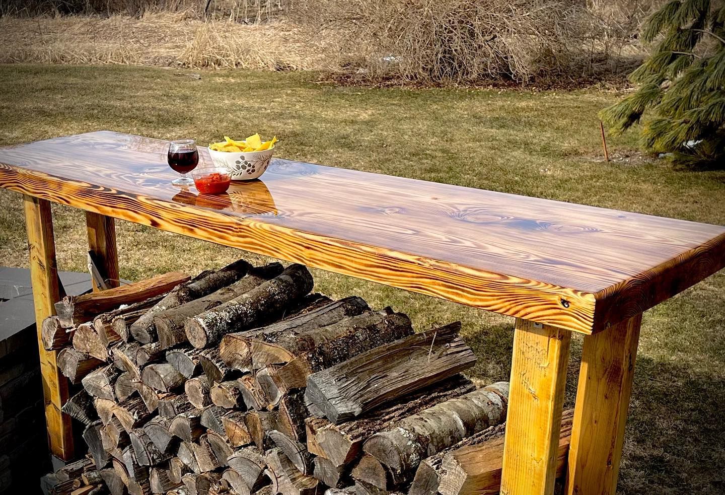 Wooden outdoor bar table with a stack of firewood below, holding snacks and a drink.