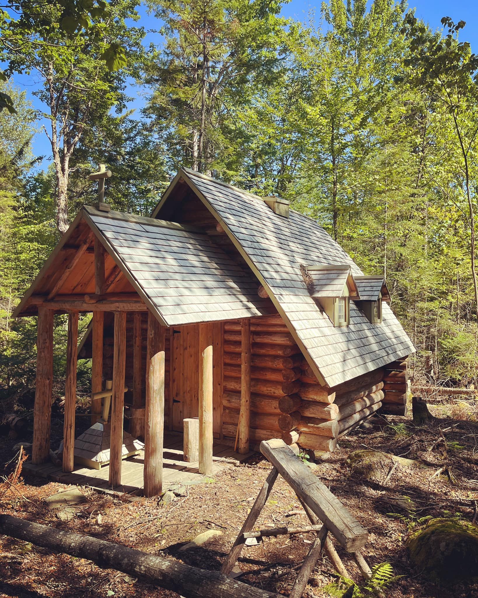 Log cabin nestled in a forest, with a porch, weathered roof, and bench in front.