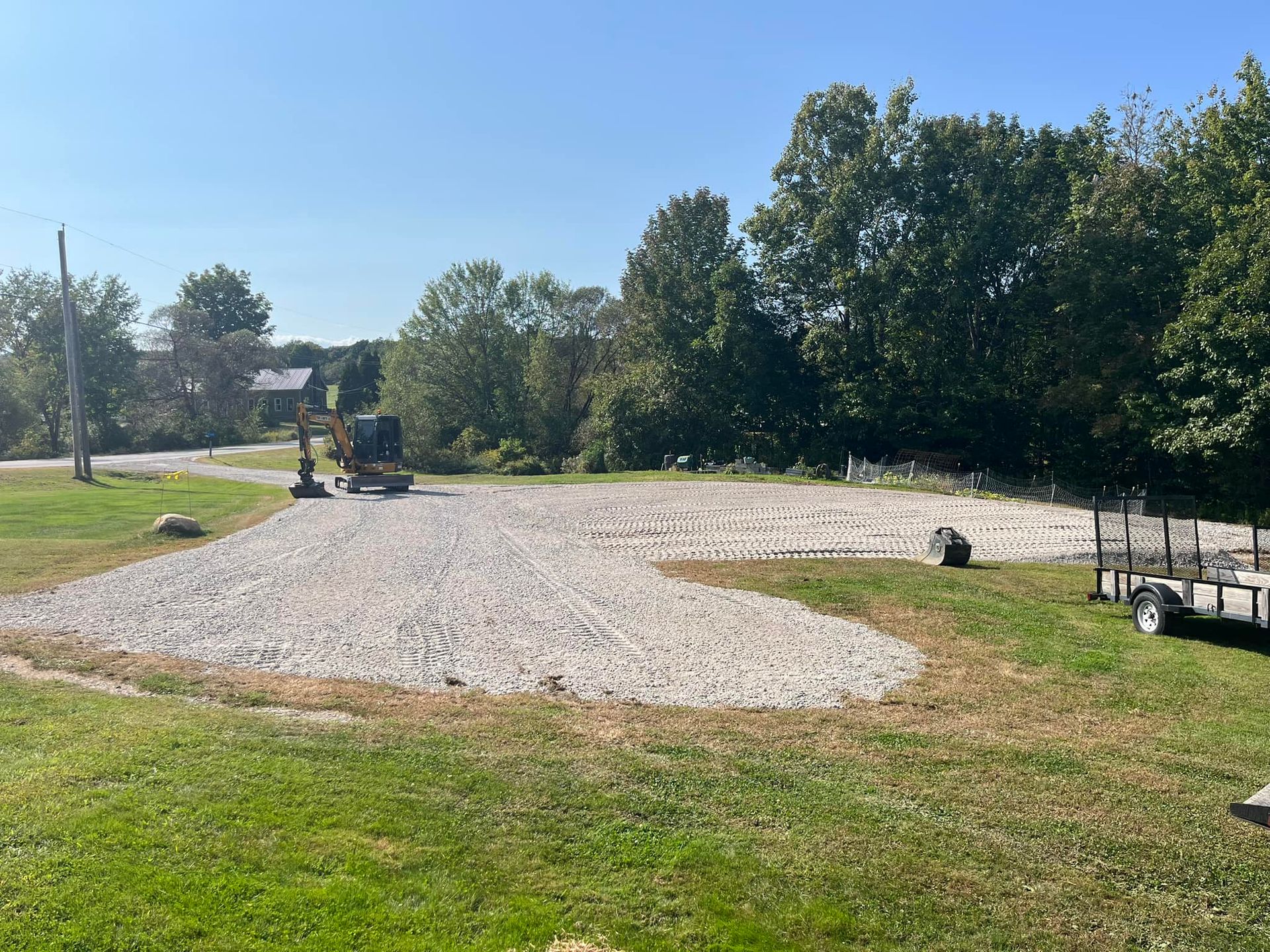 Gravel parking area under construction with an excavator and trailer on a grassy field surrounded by trees.