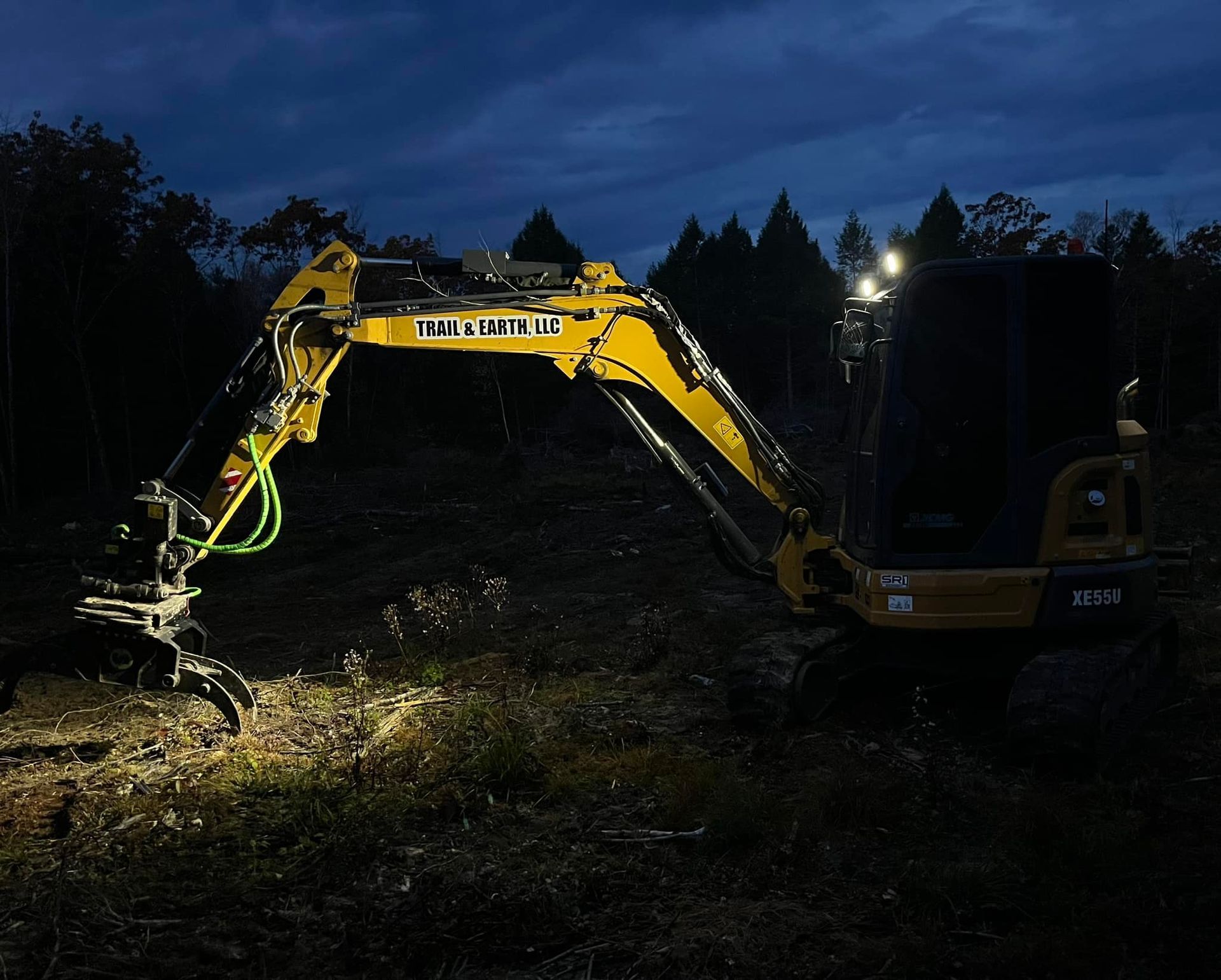 Yellow excavator with lights working in a dark forest at night.