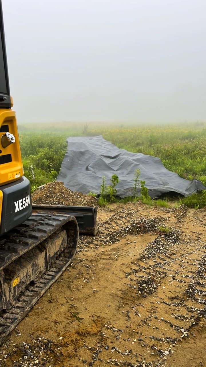 Yellow excavator on dirt, pile of gravel, black tarp in foggy field.