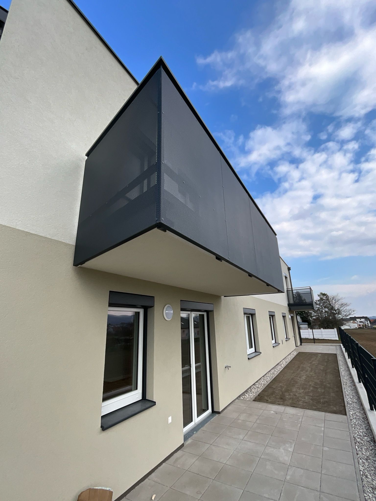 Moderne beige Gebäudefassade mit dunklem Metallbalkon, Glastüren, Fenstern und einer gepflasterten Terrasse unter blauem Himmel.