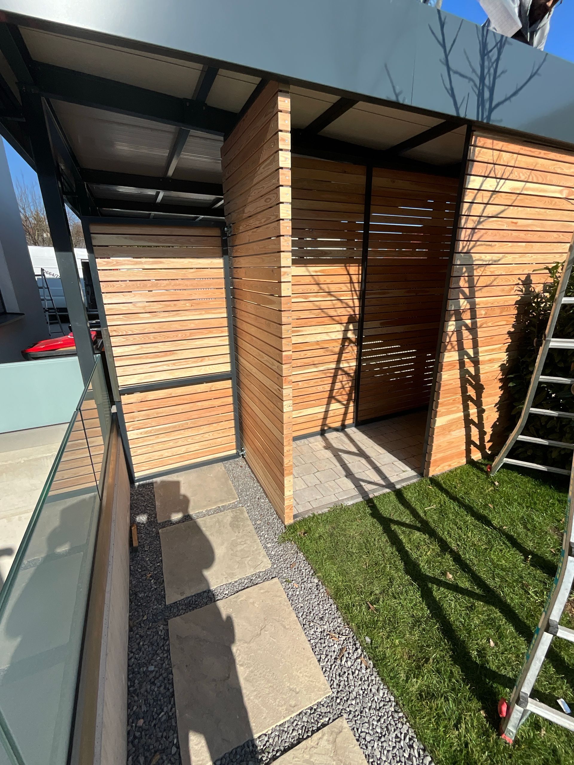 Ein modernes Carport mit einer hölzernen Lattenkonstruktion, einem Steinweg und einer kleinen Rasenfläche unter einem Metalldach.