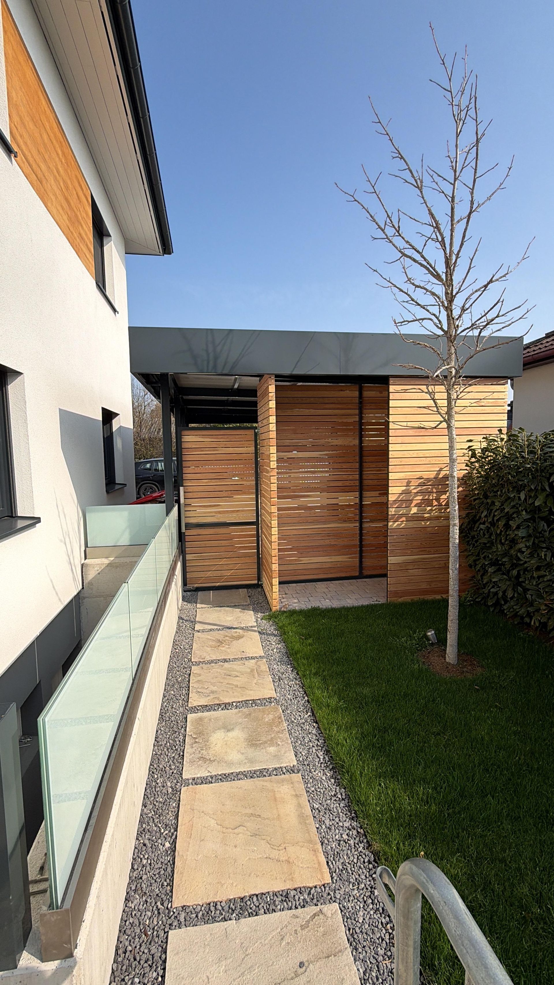 Ein Steinweg zu einer Holzkonstruktion in einem Hinterhof neben einem Haus mit einem Carport mit Glasgeländer.