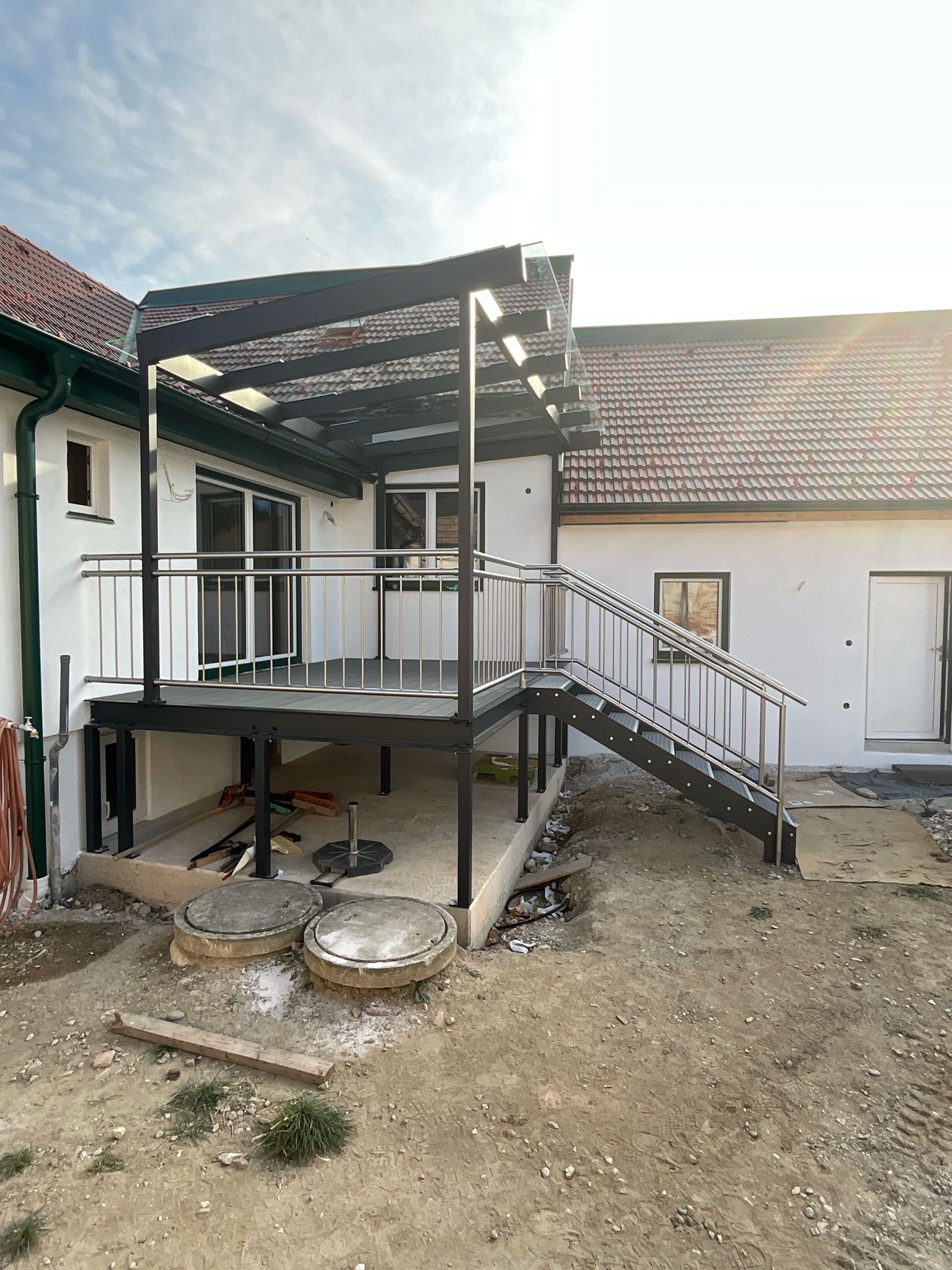 Eine neu errichtete, schwarze Metallterrasse mit Treppe und Vordach, die an der Rückseite eines weißen Hauses angebaut ist.