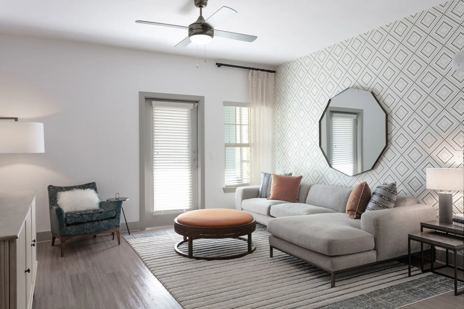 Living room with a light gray sectional sofa, orange round ottoman, and geometric wallpaper.