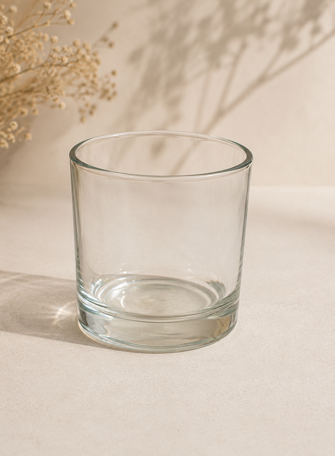 Vaso de cristal transparente sobre una superficie beige con flores secas al fondo.