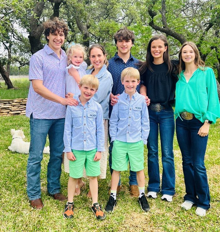 A family poses for a photo outdoors in a grassy field with trees in the background, smiling and dressed in casual attire.