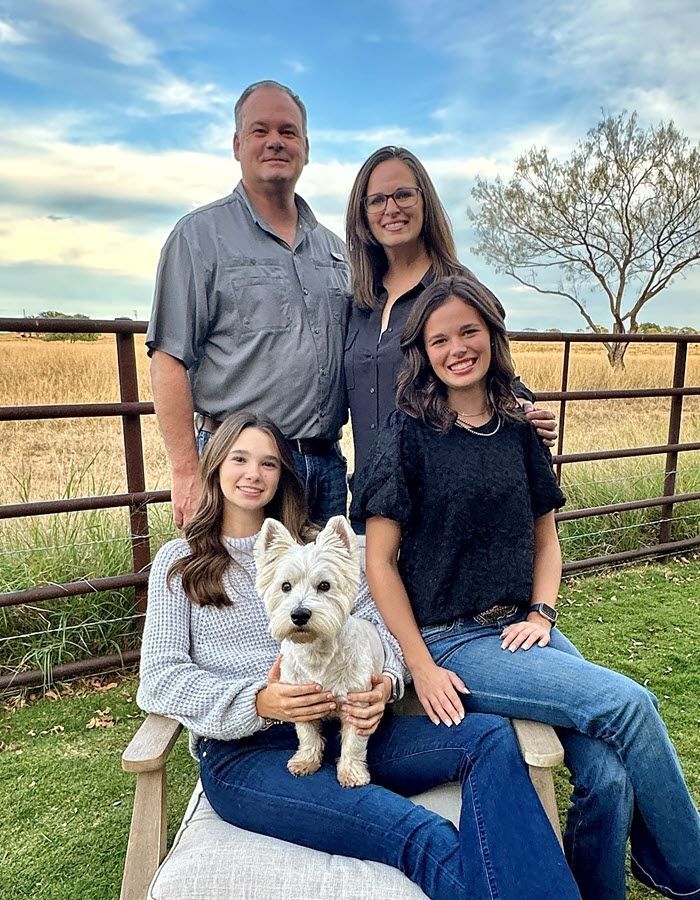 A family of four poses with their small white dog in front of a rural fence and open field under a blue sky.