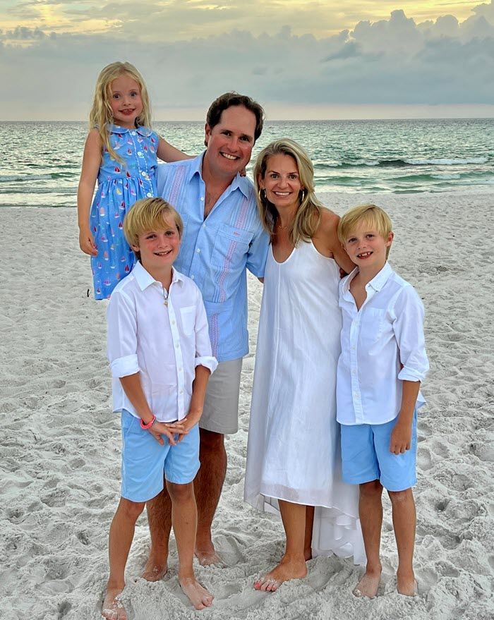 A family of five posing on a sandy beach at sunset, wearing coordinated light blue and white clothing.