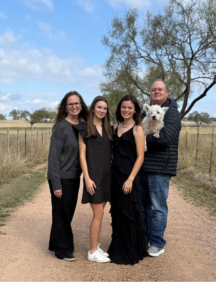 A family poses on a gravel path in a rural setting, including a small white dog held by a person in a jacket.