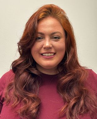 Woman with curly brown hair smiles; rosy cheeks, blue eyes, close-up shot.