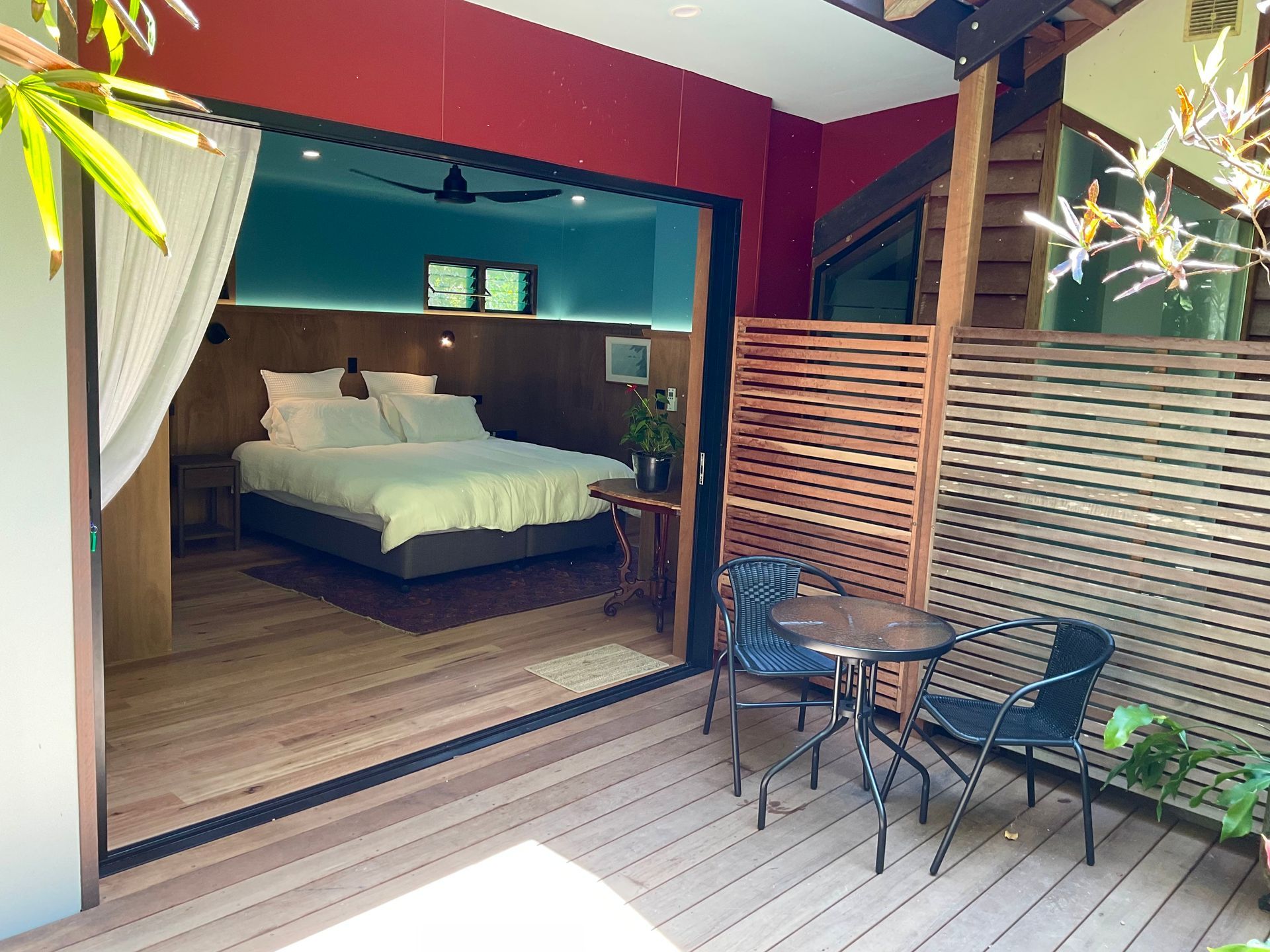 Patio opening into a bedroom with a white bed, wood deck, small table, chairs, and potted plants.