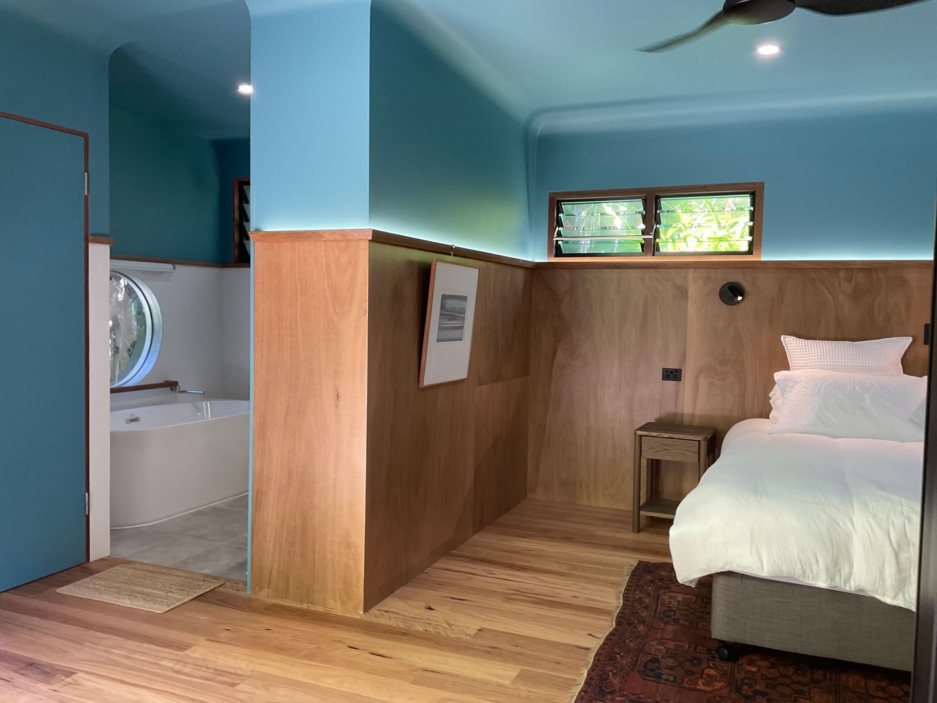 Small cozy bedroom with wood-paneled walls, bed, window, and adjoining bathroom doorway.