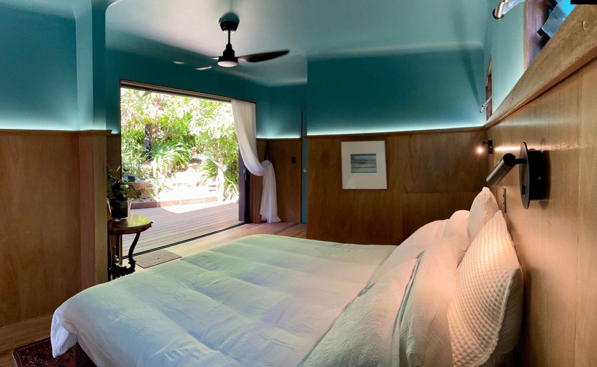 Bedroom with a large bed, teal walls, wood paneling, ceiling fan, and open sliding doors to greenery