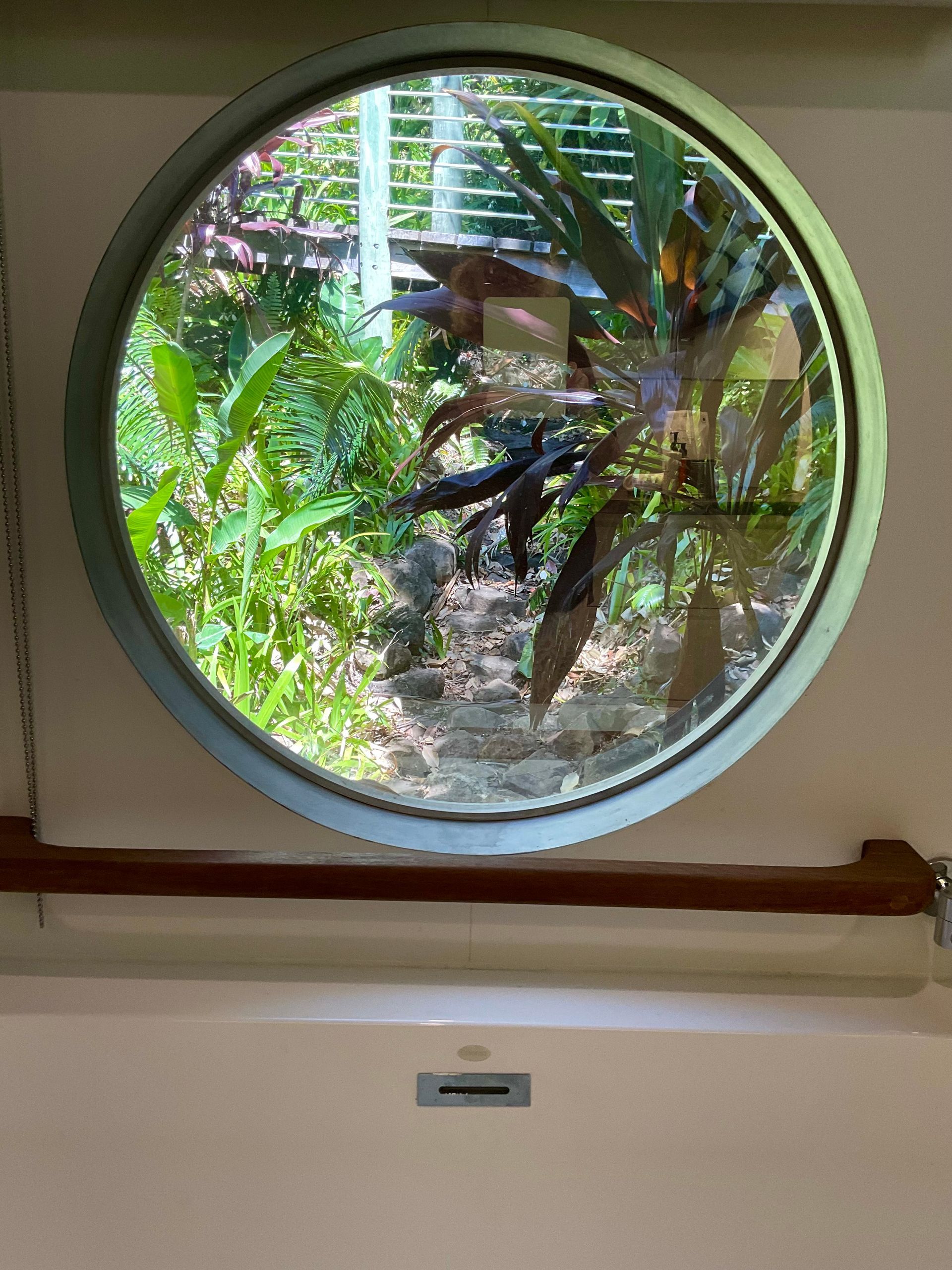Round window overlooking lush green plants and a wooden deck outside.