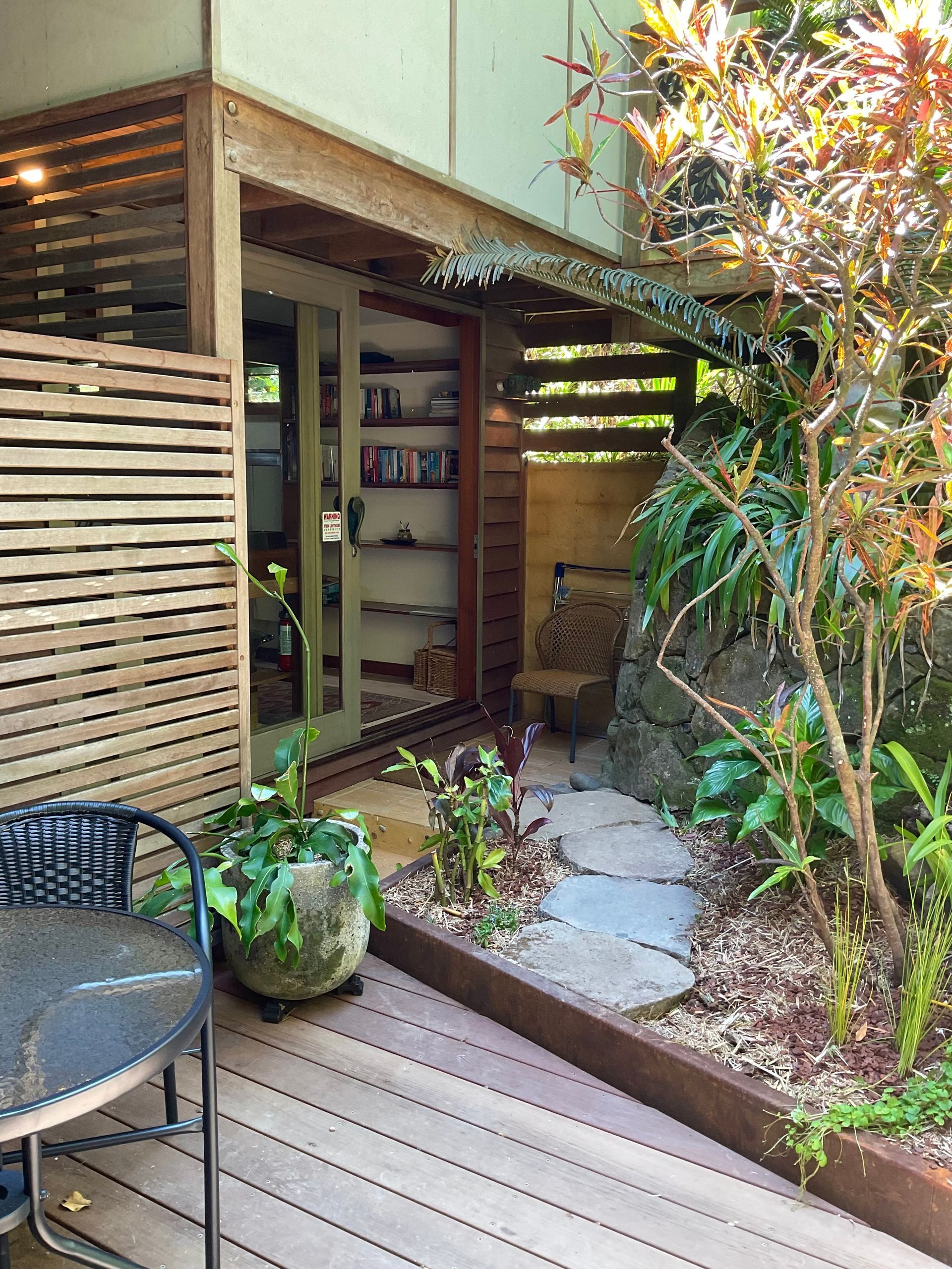 Wooden patio entrance with potted plants, a chair, and greenery beside a modern house