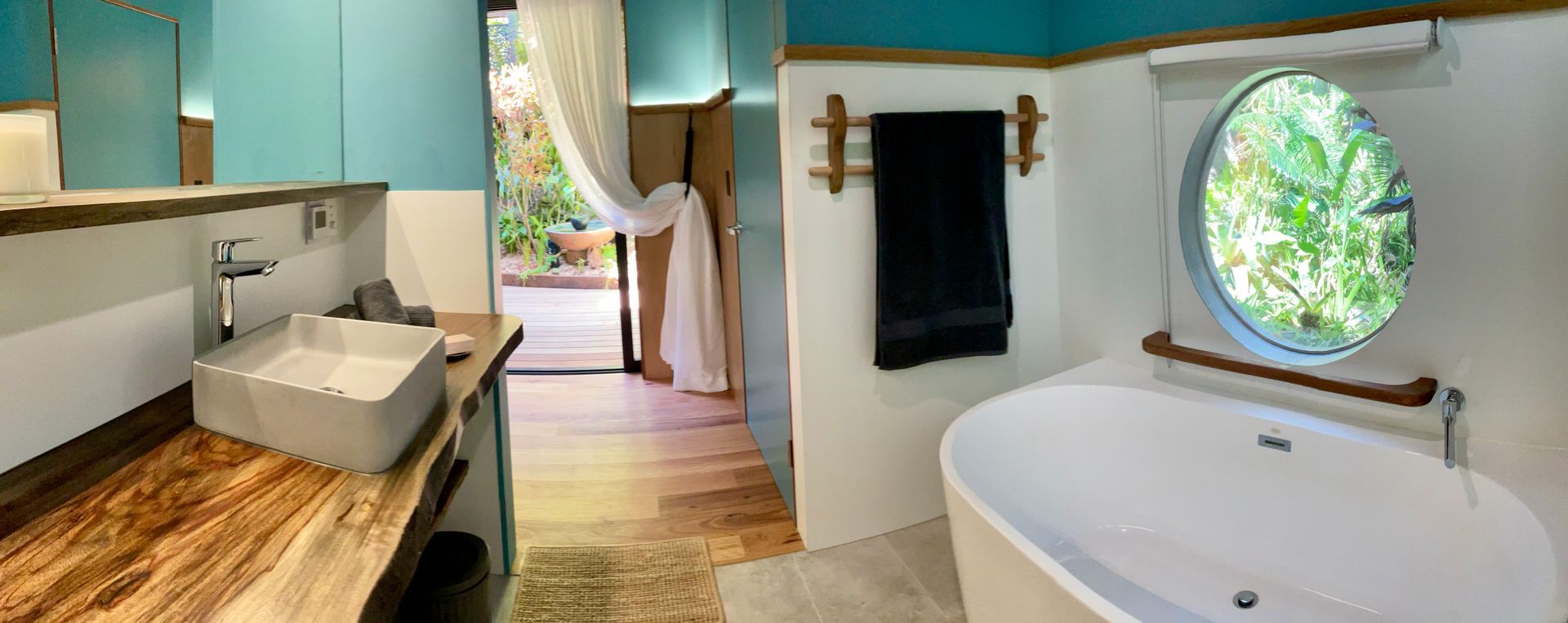 Bright bathroom with double sink, wooden counter, freestanding tub, round window, and open door to greenery