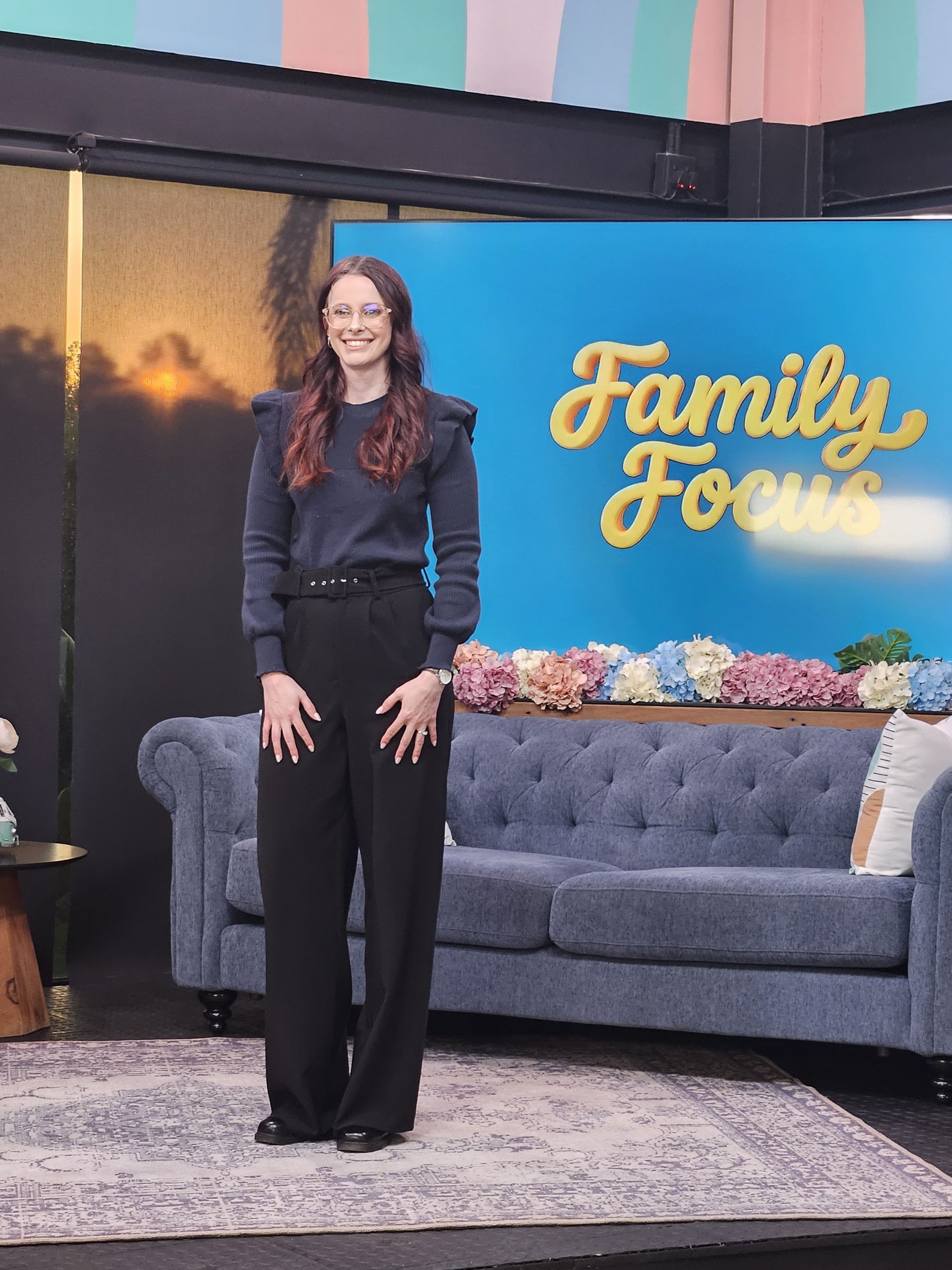 Nikki Russell standing on the SABC2 Eintlek Let's Connect studio set beside a gray couch and “Family Fours” sign