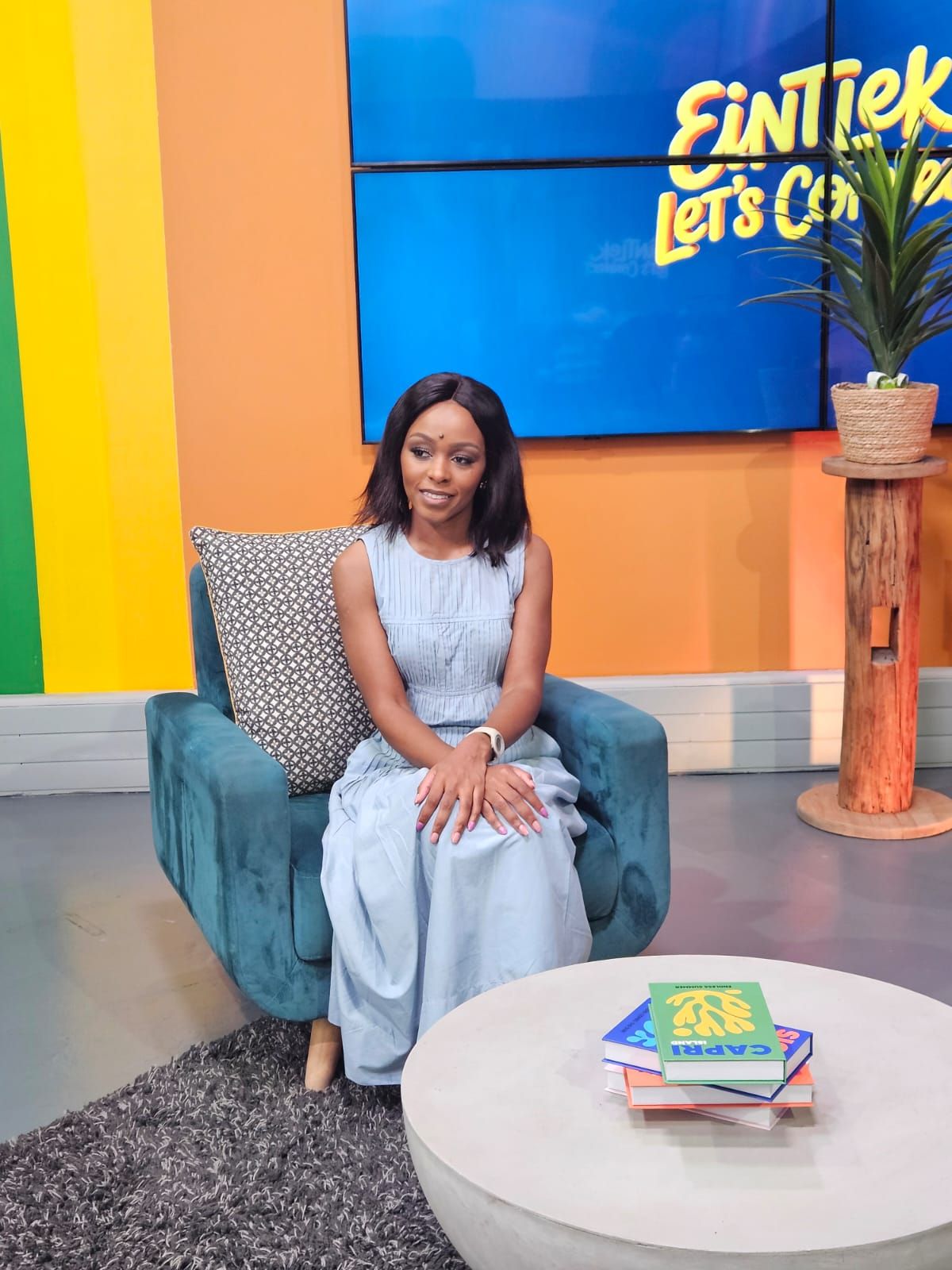 Phozisa sits in a teal armchair in a television studio with a colorful wall and a plant on a wooden pedestal nearby.