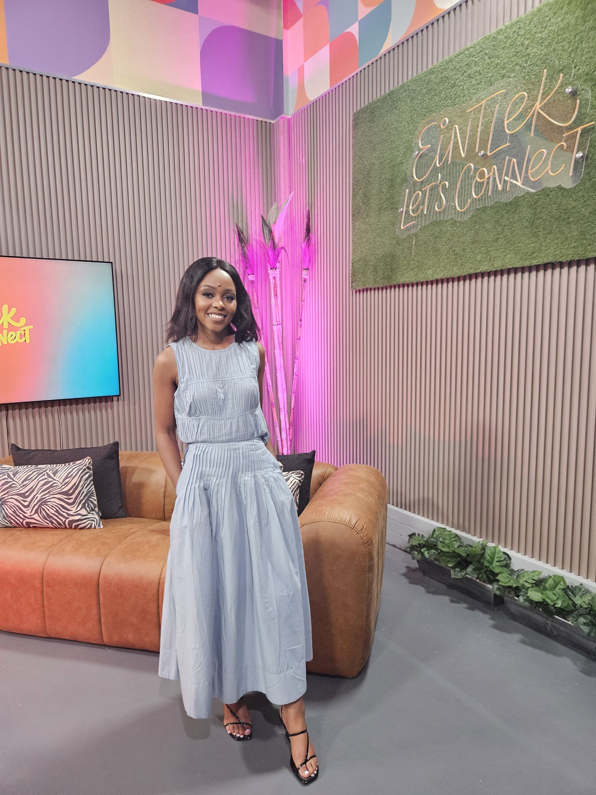 Phozisa wearing a light blue, textured sleeveless dress stands smiling in a studio with a leather sofa and green wall.
