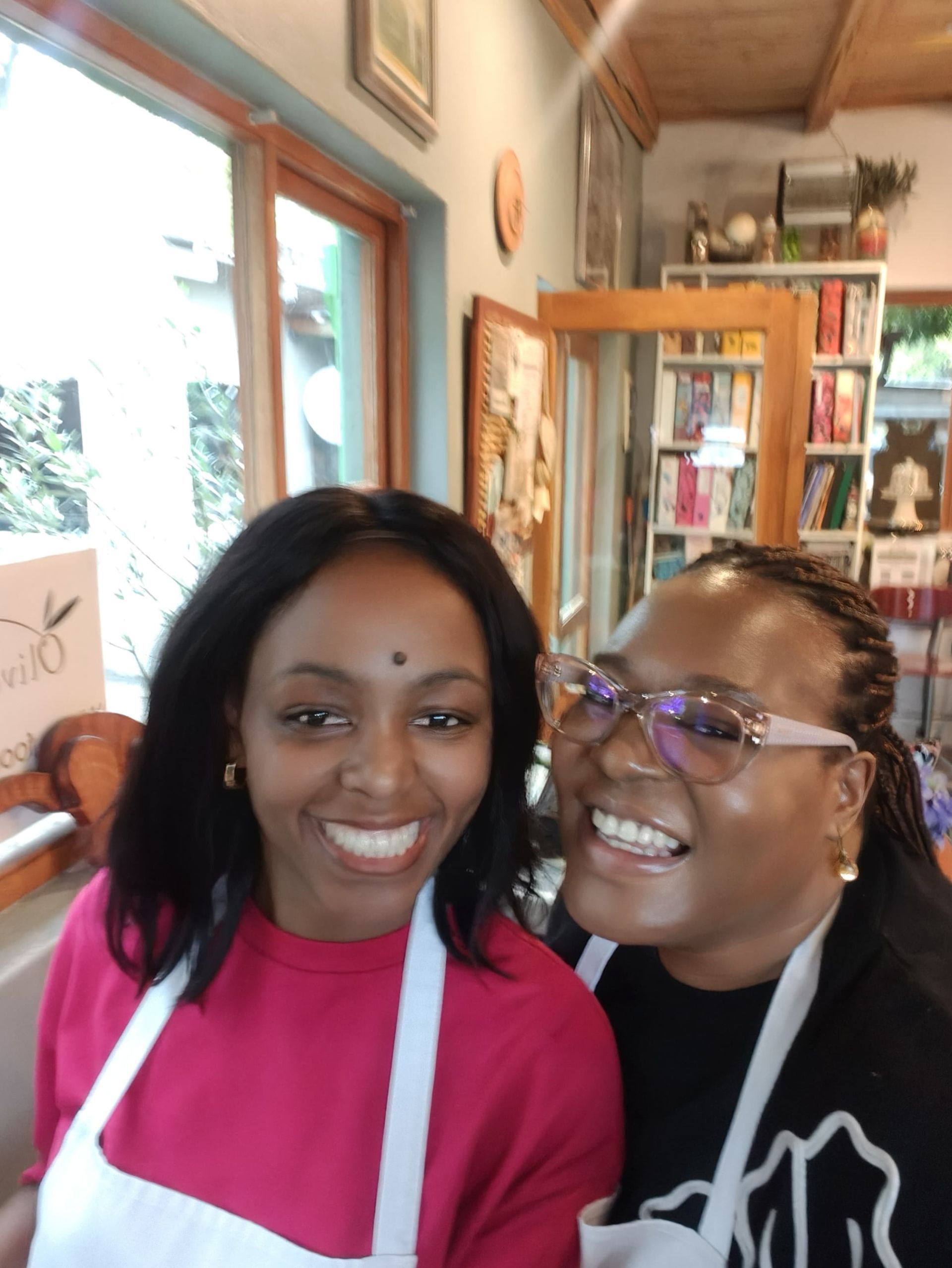 Sandy and Phozisa wearing white aprons   while standing inside a bright room with bookshelves.