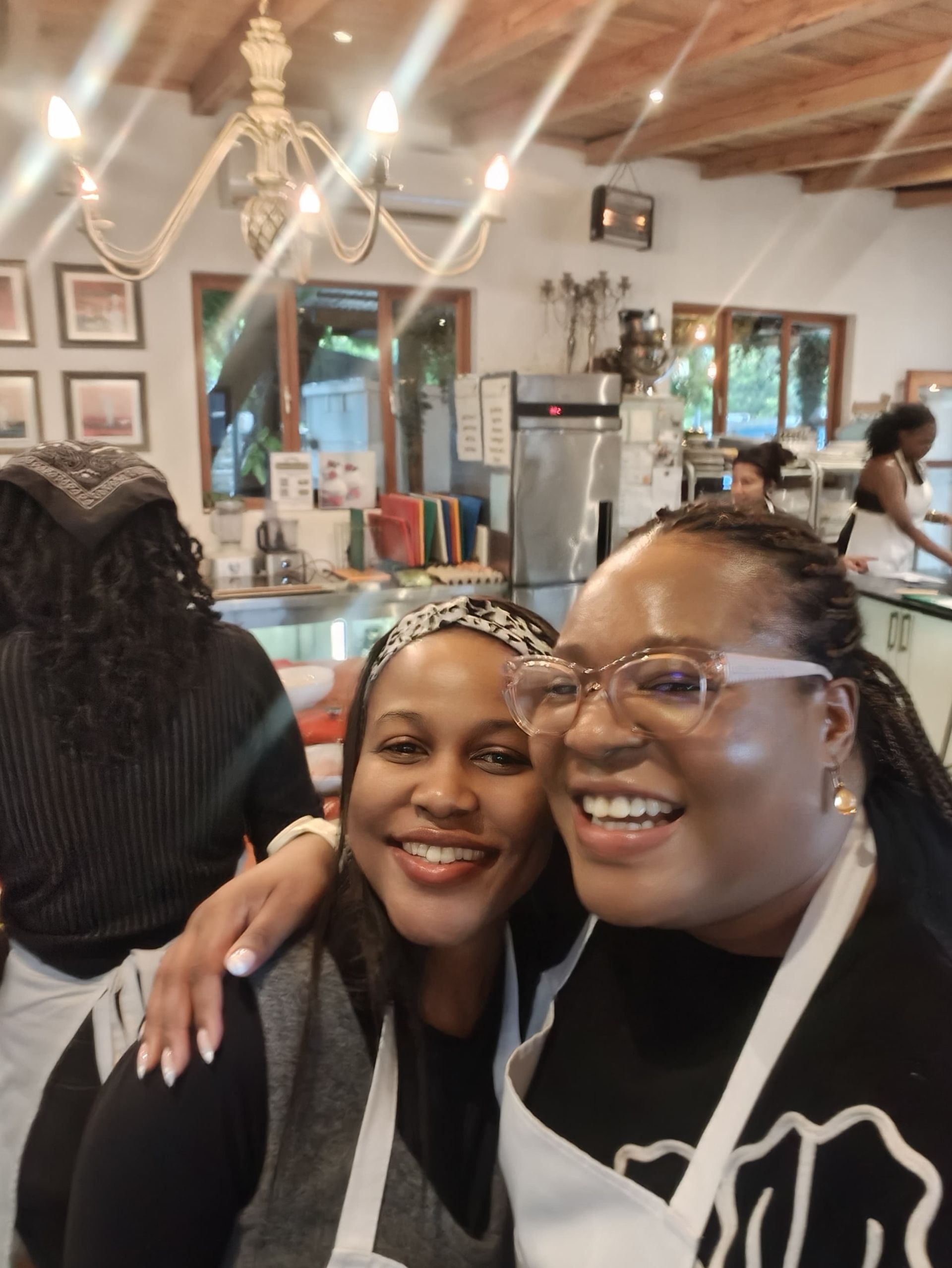 Sandy Kass and Zinzisa Deliwe -Juju smiling in aprons pose for a selfie inside a brightly lit kitchen.
