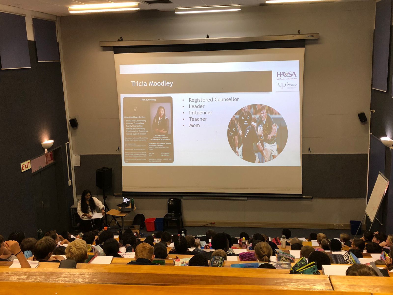 Tricia Moodley presenting on a large screen in a lecture hall with an audience seated in tiered rows.