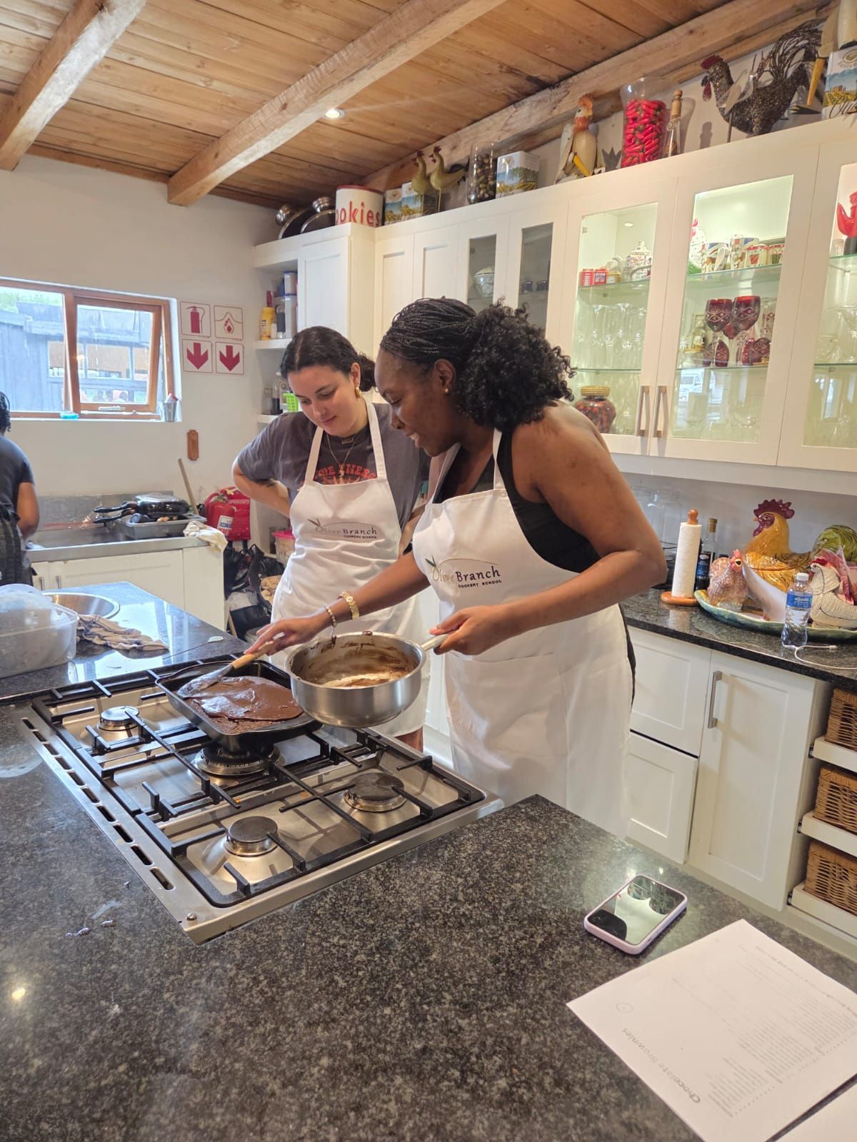 Tanna and Lucia in white aprons stand in a kitchen, cooking together on a stovetop.