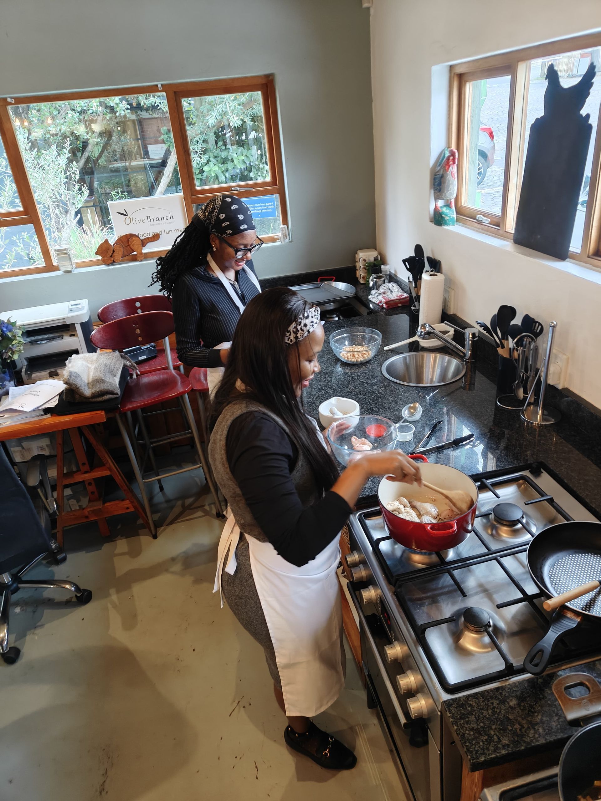 Two individuals wearing aprons stand in a kitchen, one stirring a red pot on the stove while the other looks on.