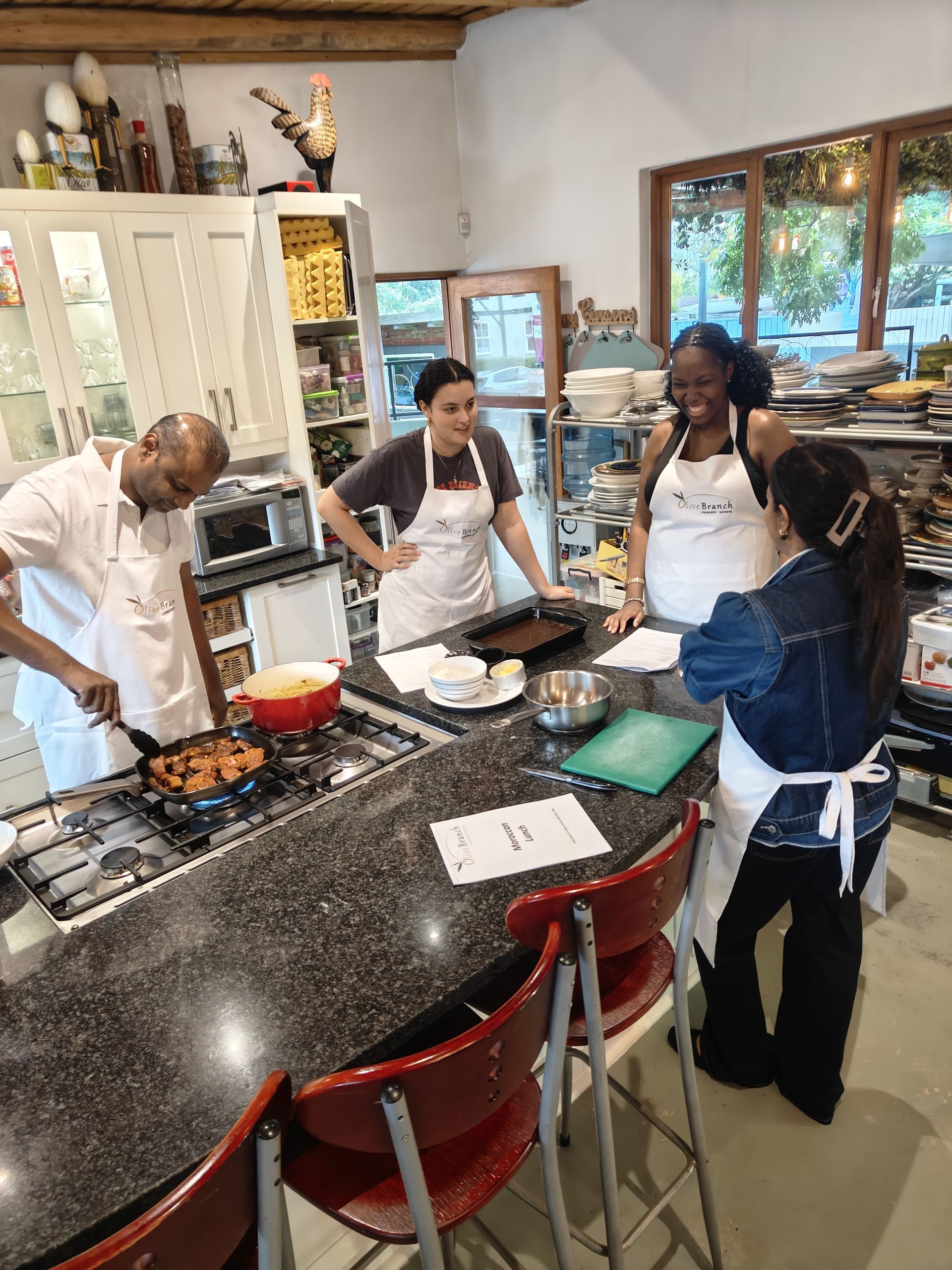 Tanna Beautement and Lucia wearing white aprons gather around a kitchen.