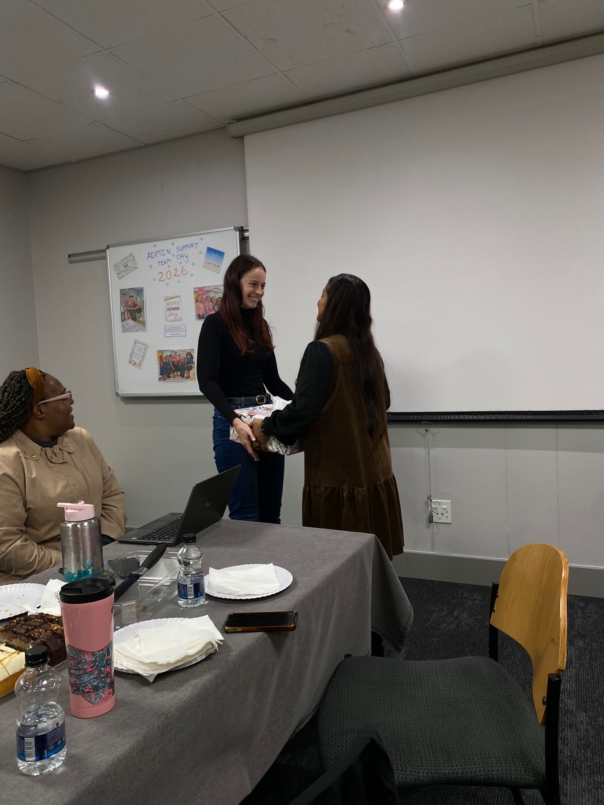 Nikki Russell and Tricia Moodley shaking hands in a classroom with a projector screen and table in the foreground.