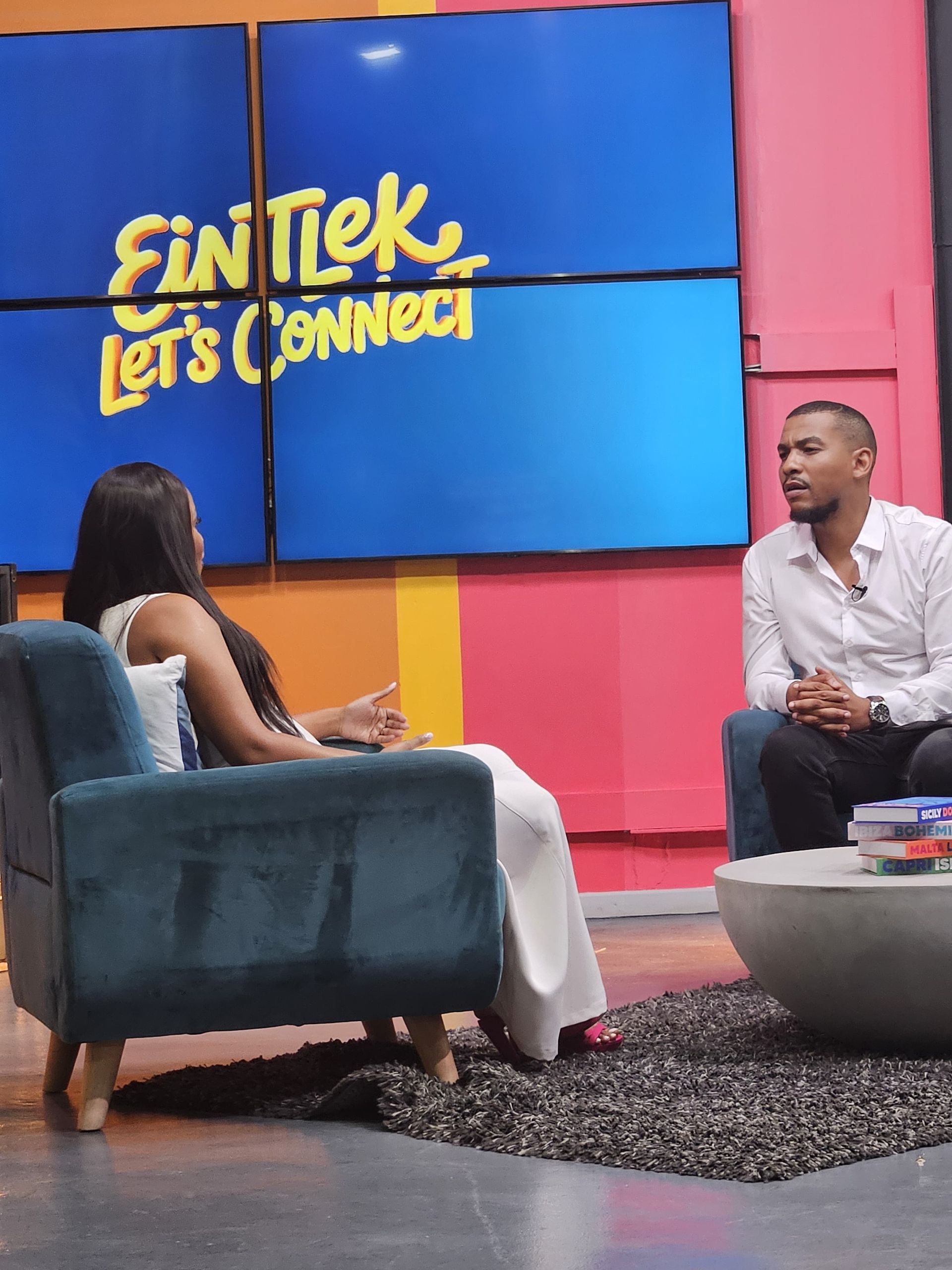 Fran-Rico and Zinzisa sit in armchairs on a colorful set, having a conversation in front of a screen reading 