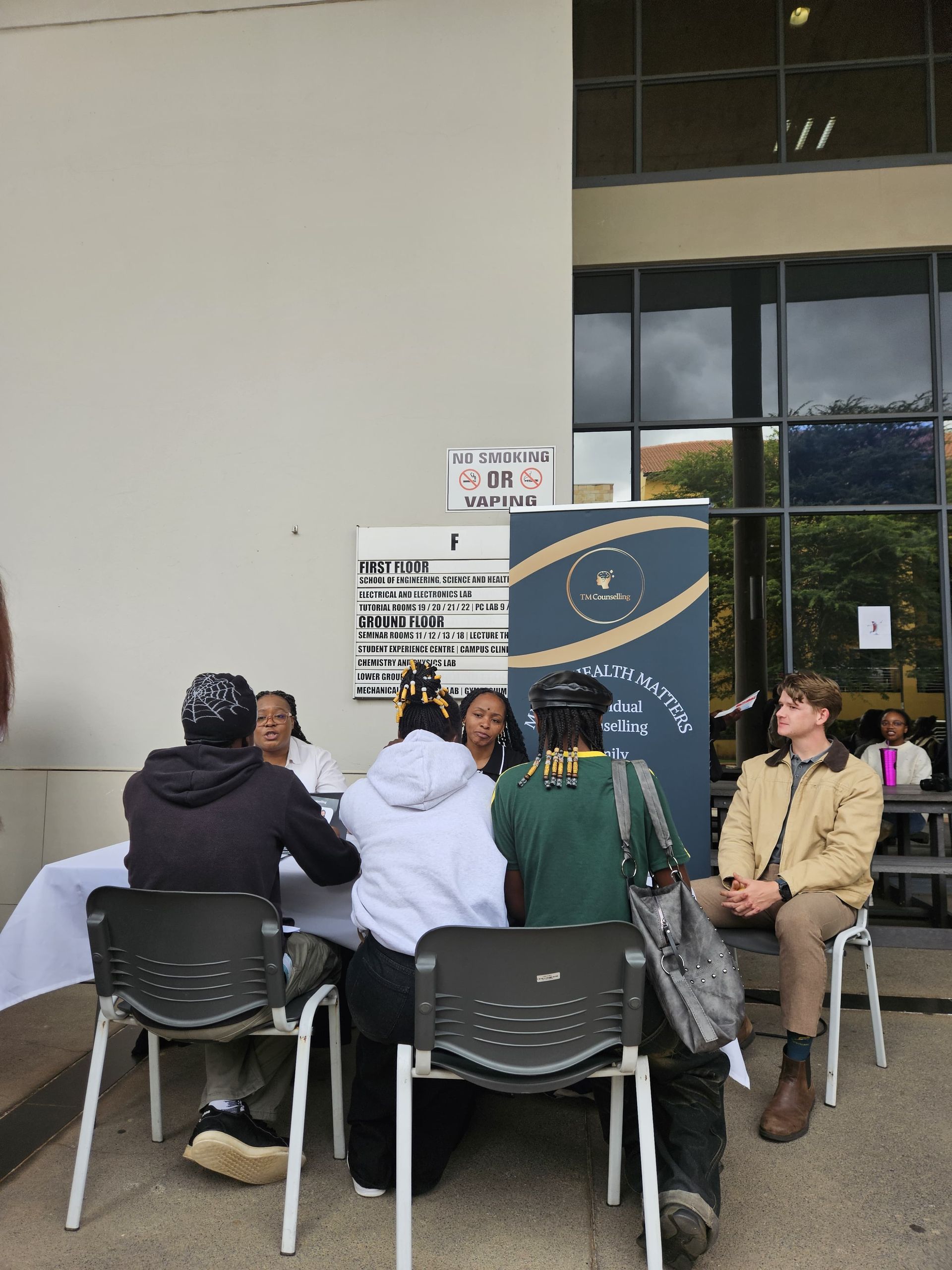 Five people sit at a table outdoors in front of a blue informational sign, engaging in a conversation or meeting.