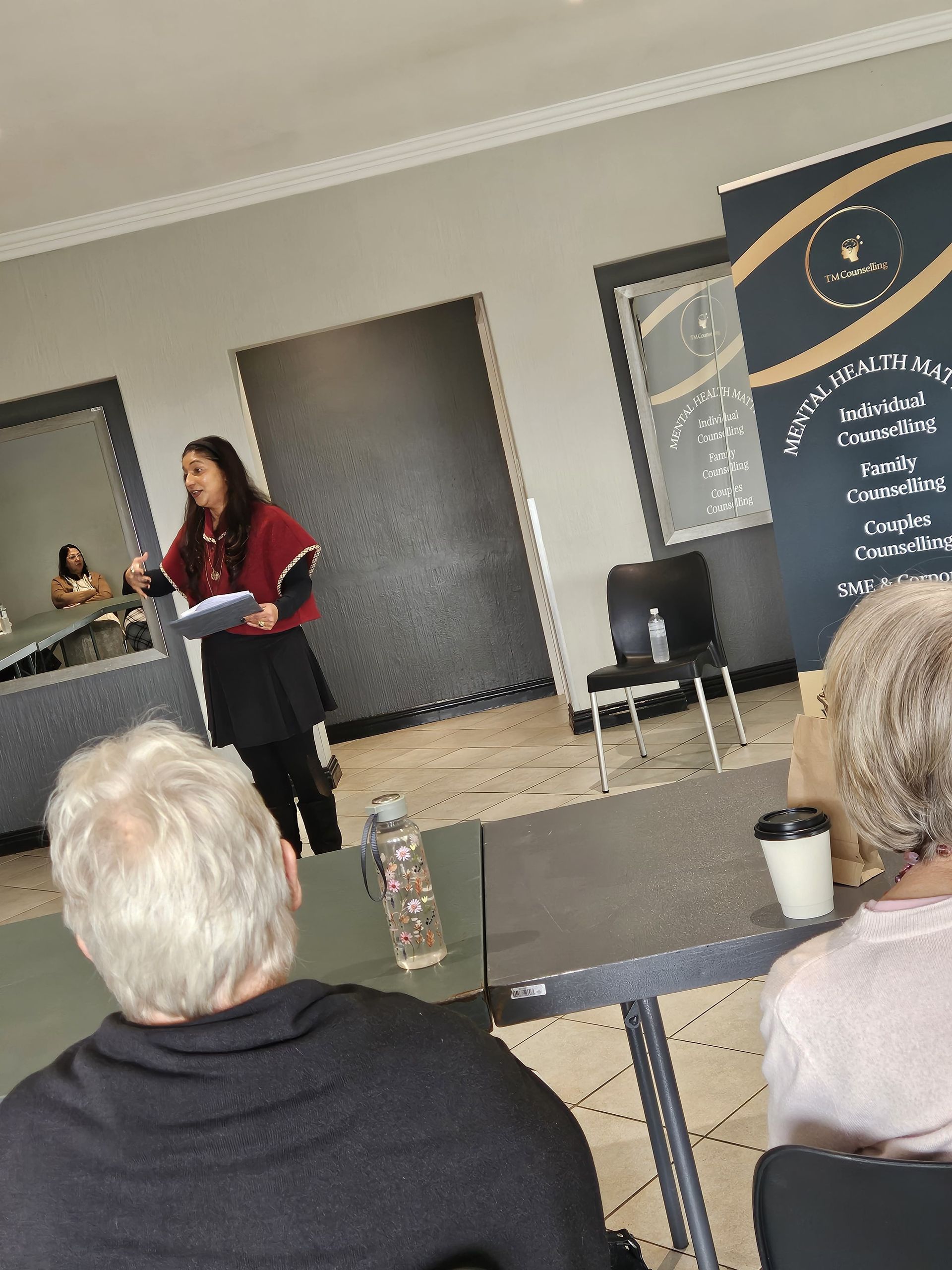 Tricia Moodley stands presenting to an audience in a room, with a tall branded banner positioned to the right.