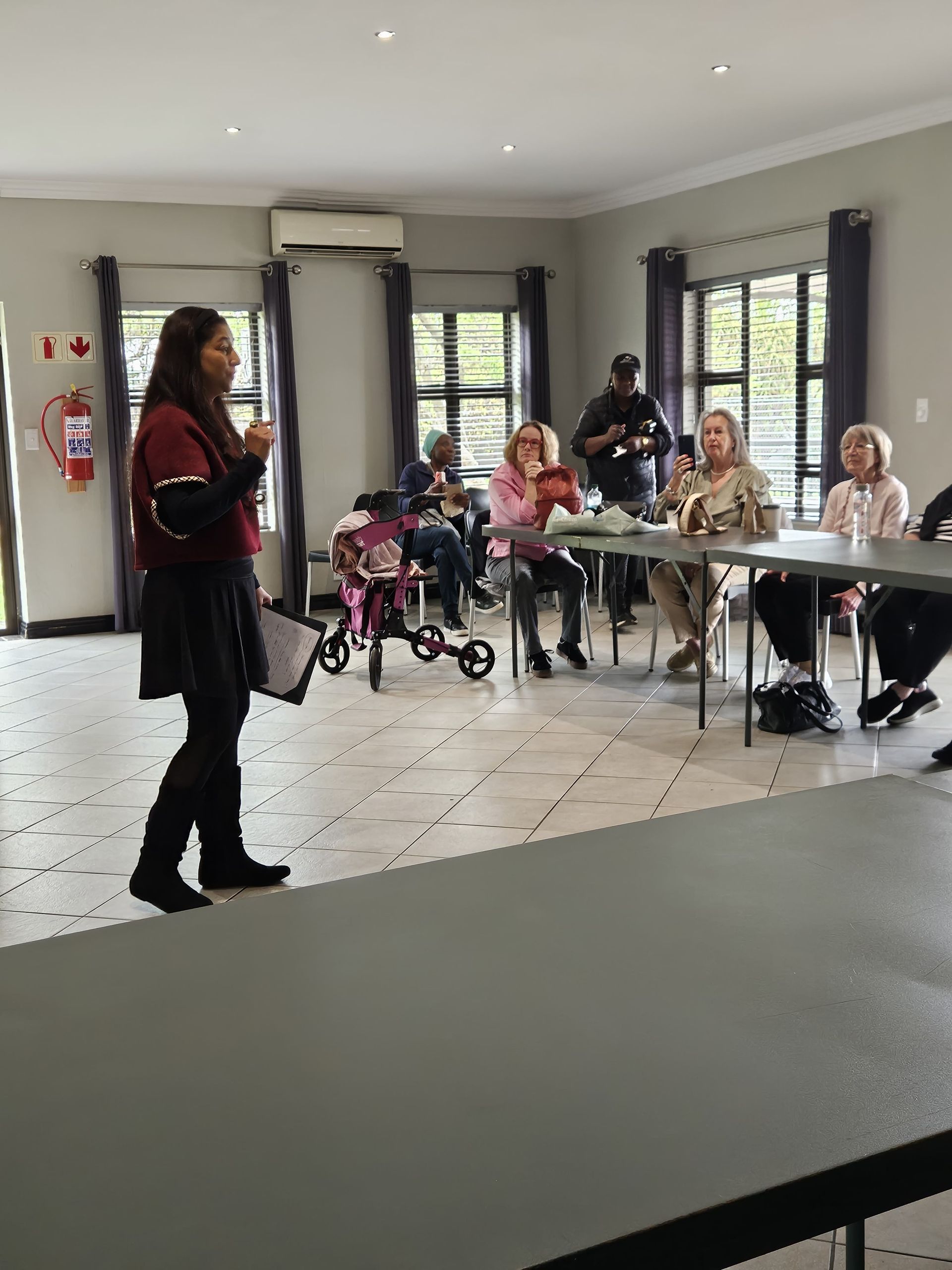 Tricia Moodley stands in a bright room, gesturing to an audience seated at tables during a meeting or presentation.